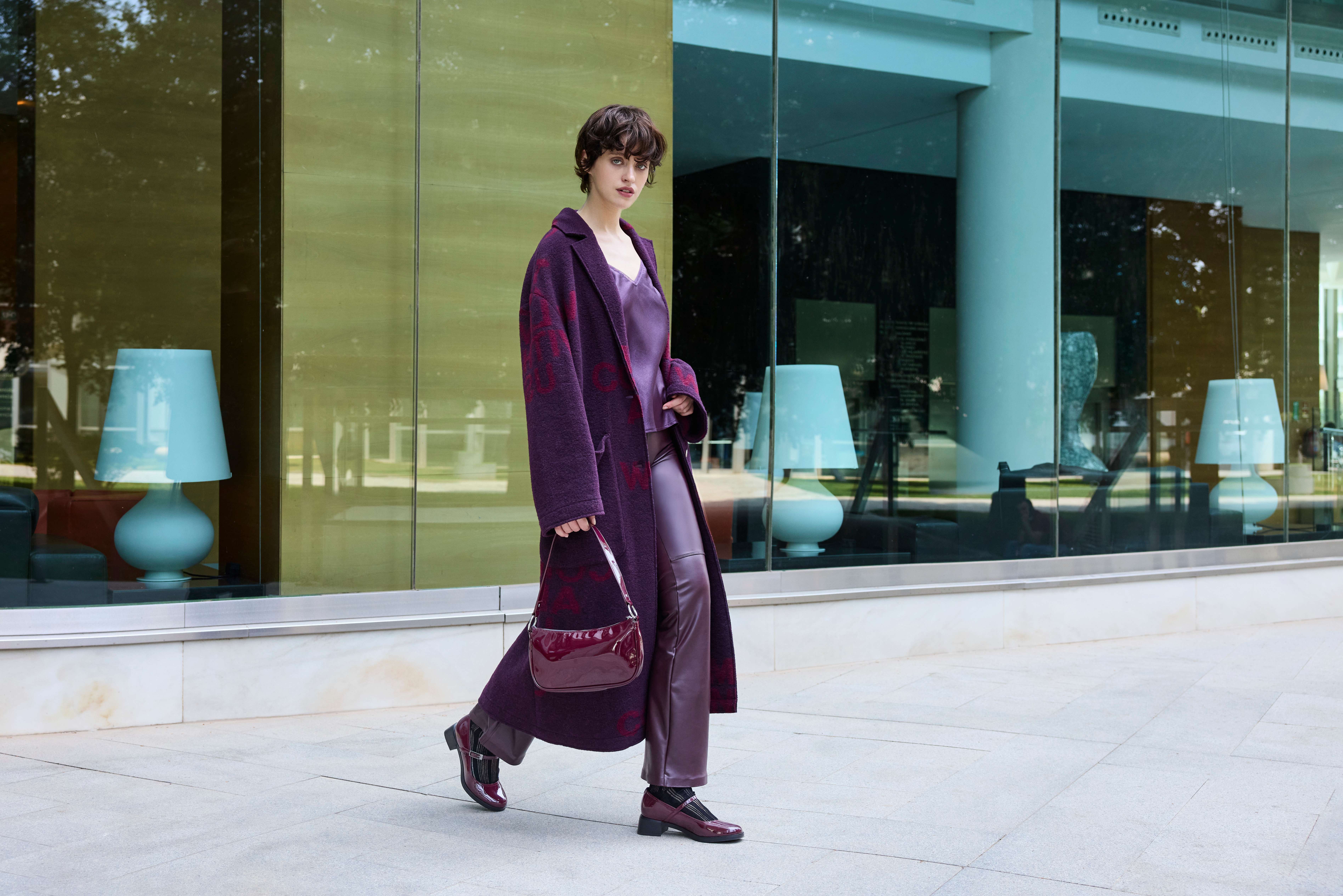 Una donna indossa un cappotto bordeaux con pantaloni lucidi, un top abbinato, una borsetta e scarpe di vernice
