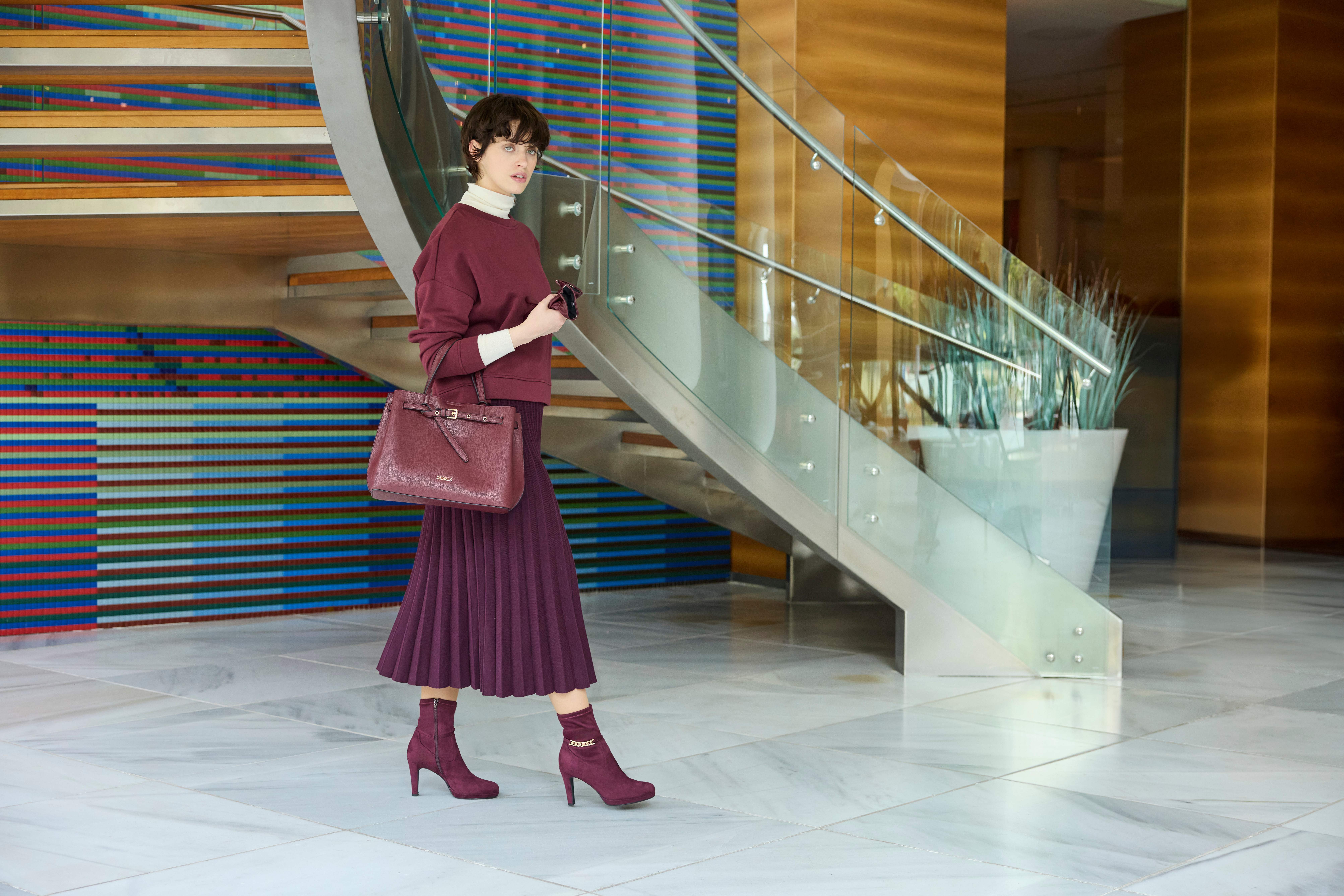 Una donna indossa un maglione bordeaux sopra un dolcevita beige, una gonna a pieghe abbinata, stivaletti e una borsa capiente