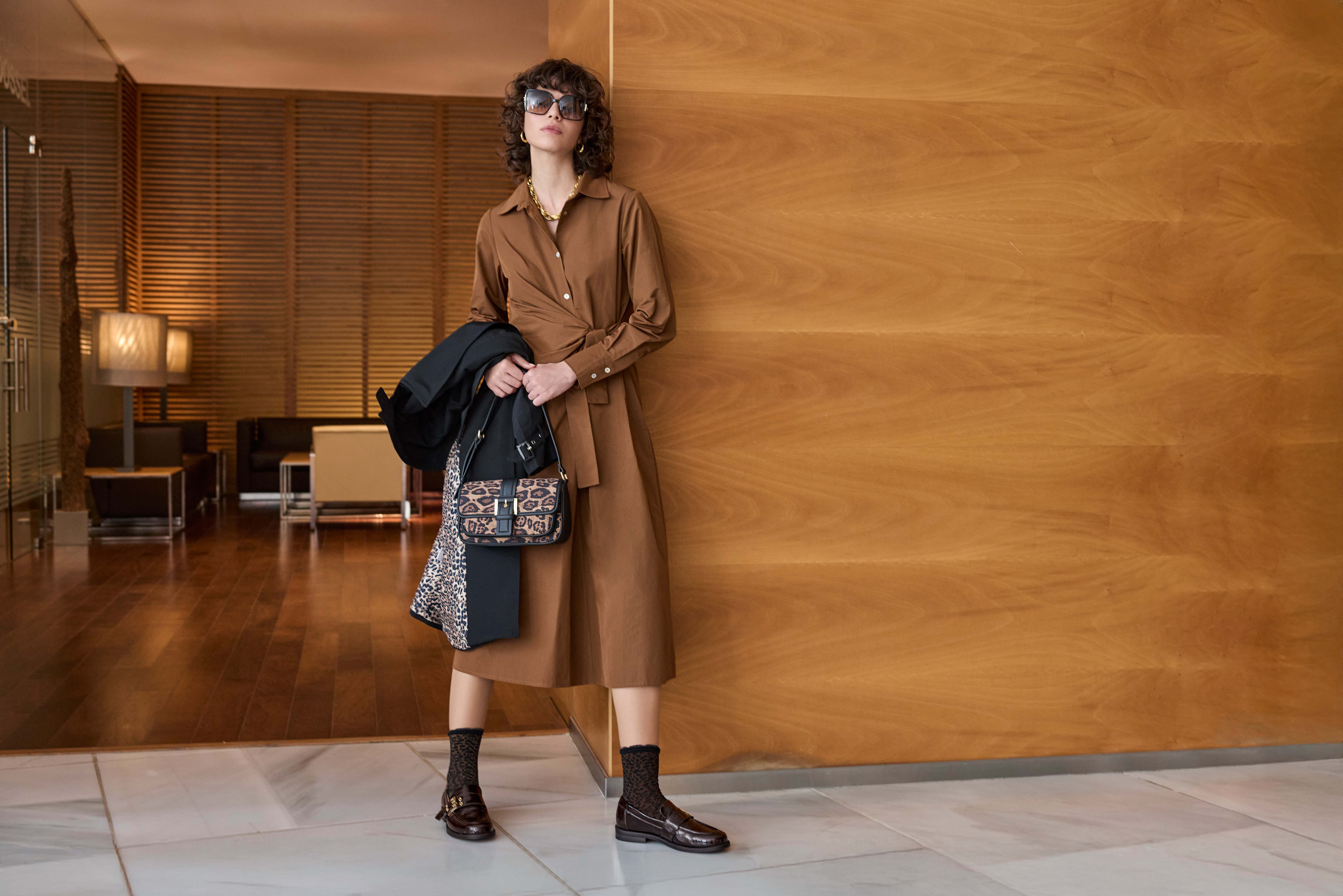 Eine Frau trägt ein braunes Hemdblusenkleid mit Loafern, Statement-Tasche und Sonnenbrille.