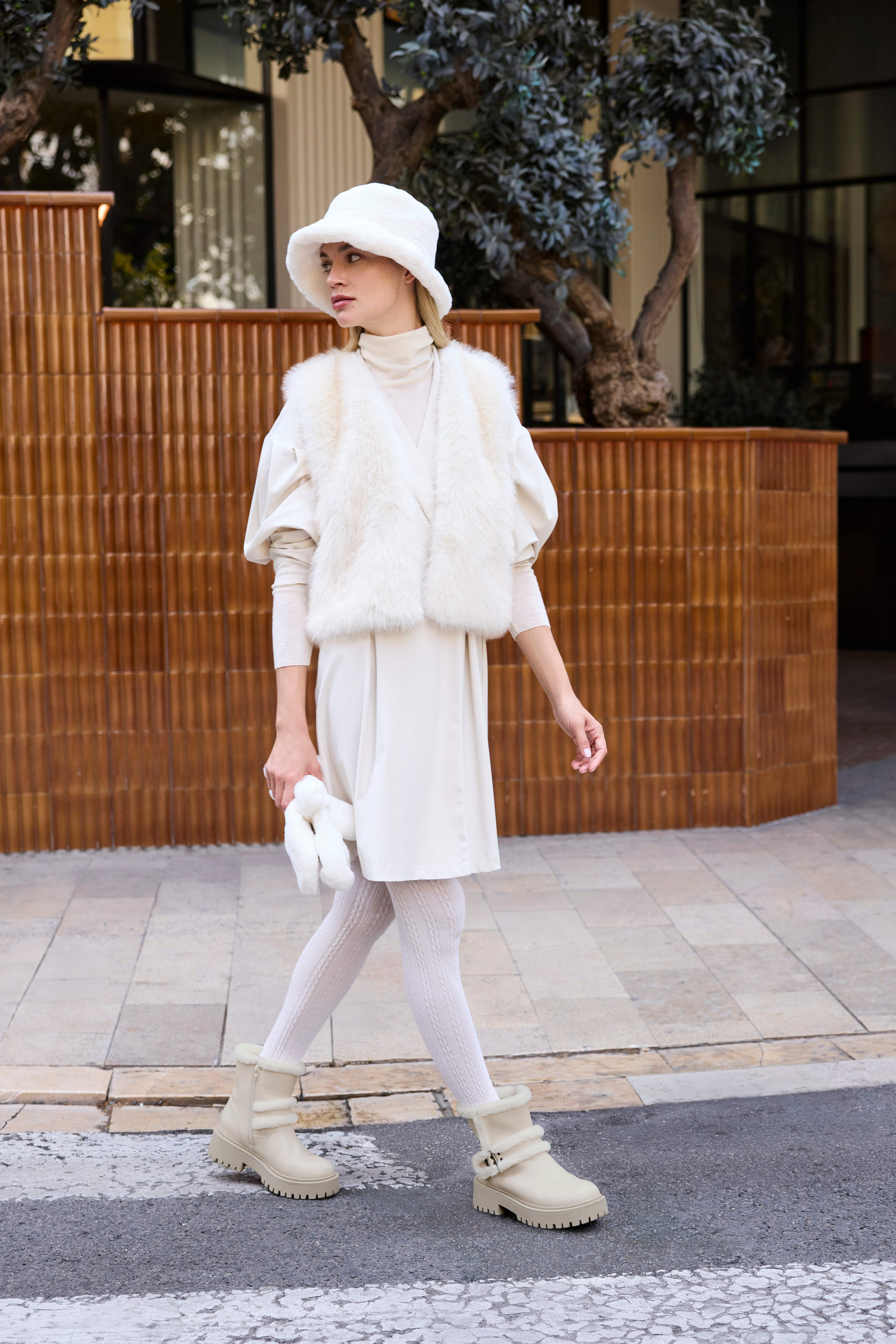 Eine Frau trägt ein komplett cremefarbenes Outfit mit Kleid, Strickstrumpfhose, Fellweste, Hut und passenden Boots