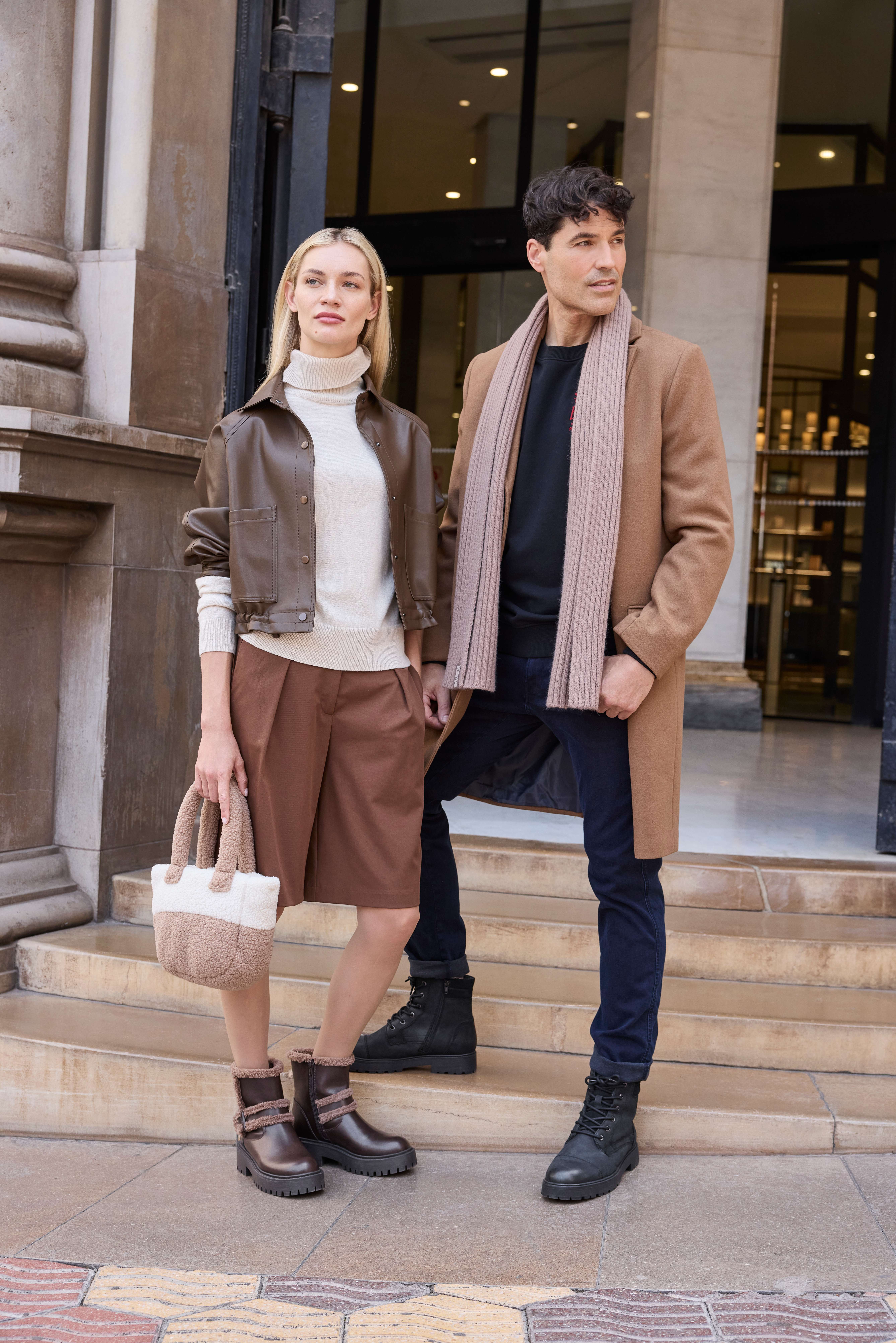 Eine Frau trägt ein Outfit aus Lederjacke, Rollkragen und Shorts; ein Mann kombiniert Mantel, Schal und Boots.