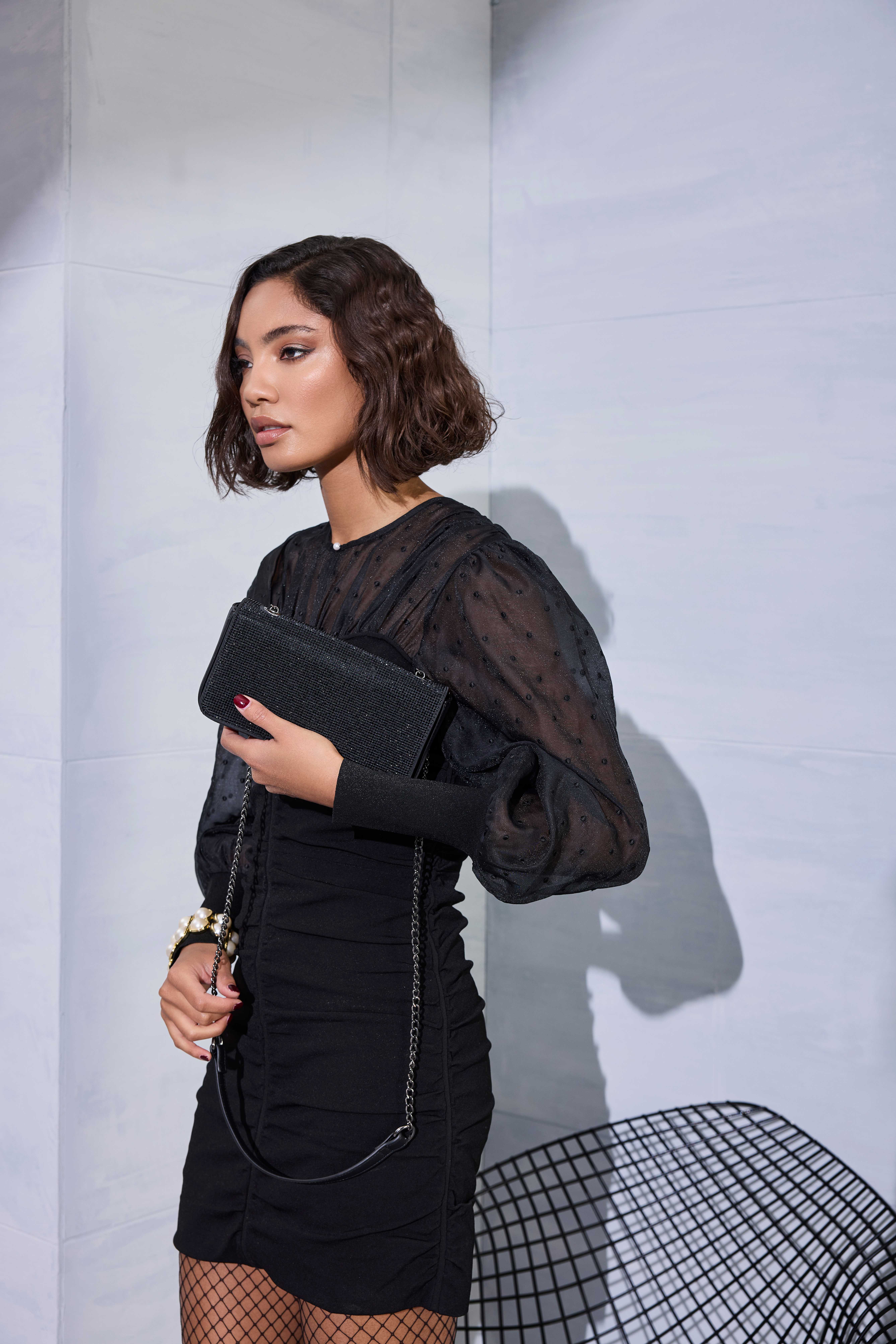 Model im schwarzen Kleid mit Transparentärmeln hält glitzernde Clutch mit Kettenriemen