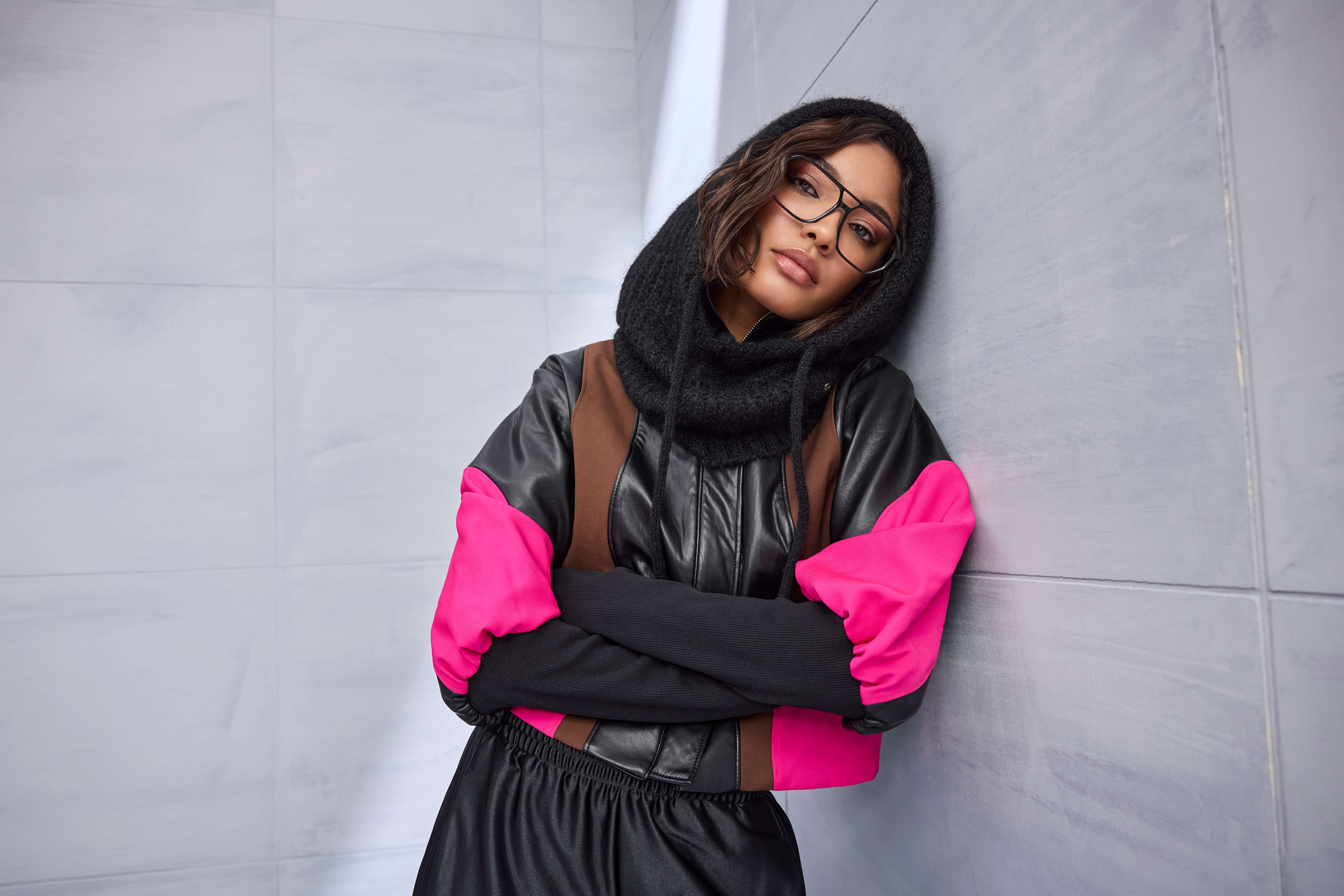 Frau in schwarzer Lederjacke mit pinken Colourblock-Ärmeln trägt gestrickte Kapuze und Brille