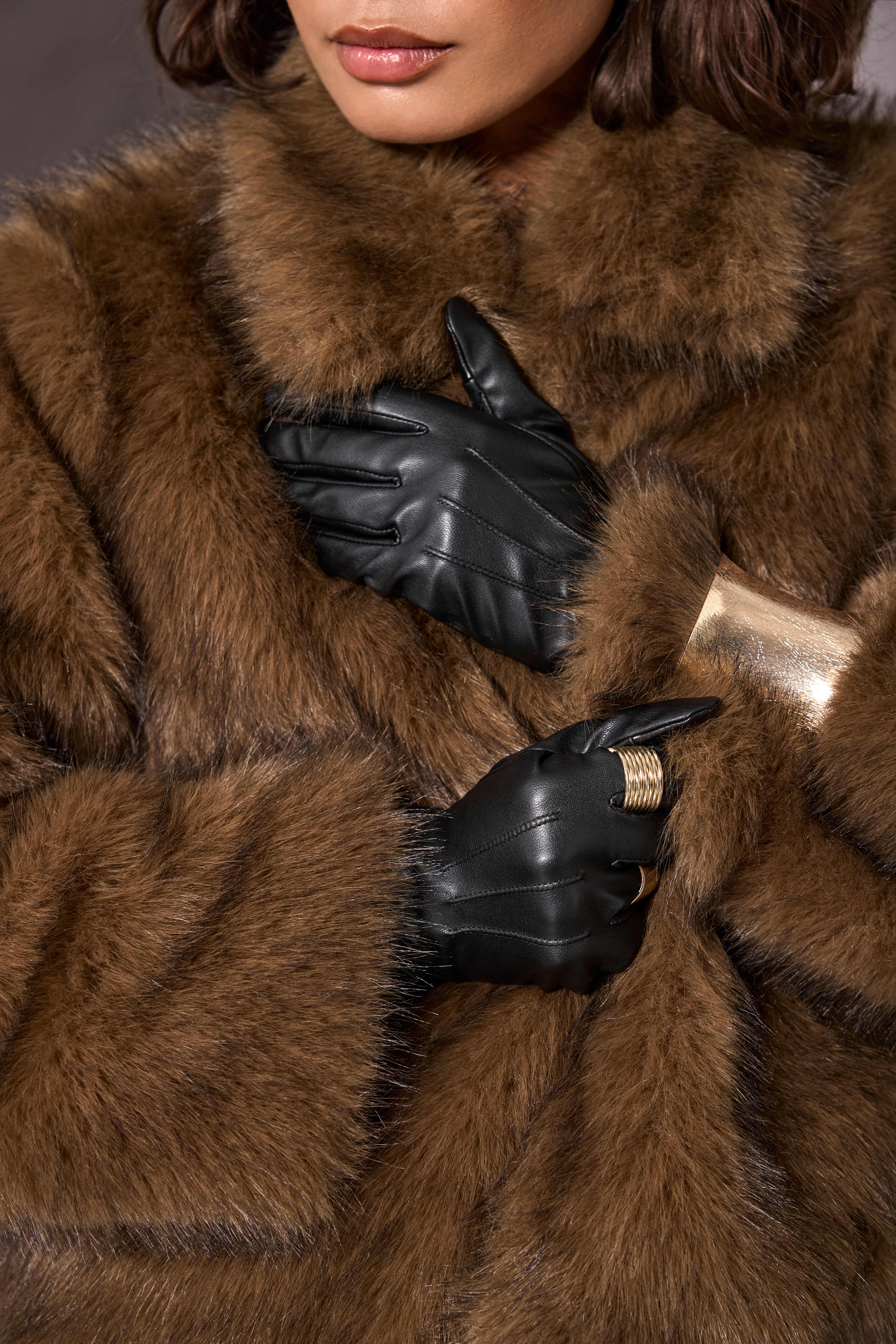 Nahaufnahme eines braunen Pelzmantels mit schwarzen Lederhandschuhen und goldenen Ringen
