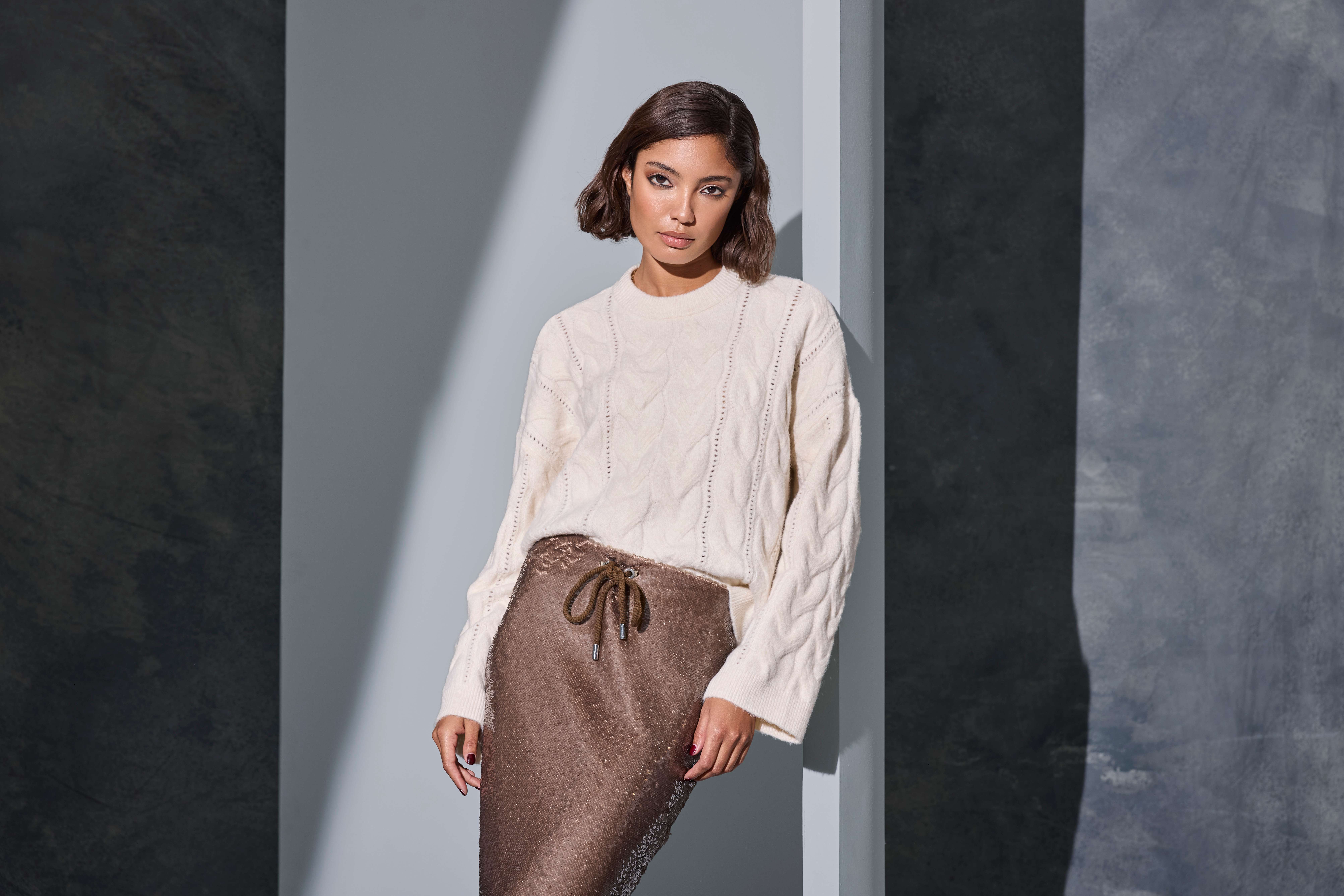 Eine Frau trägt einen cremefarbenen Strickpullover und einen braunen Rock vor einer modernen Wand