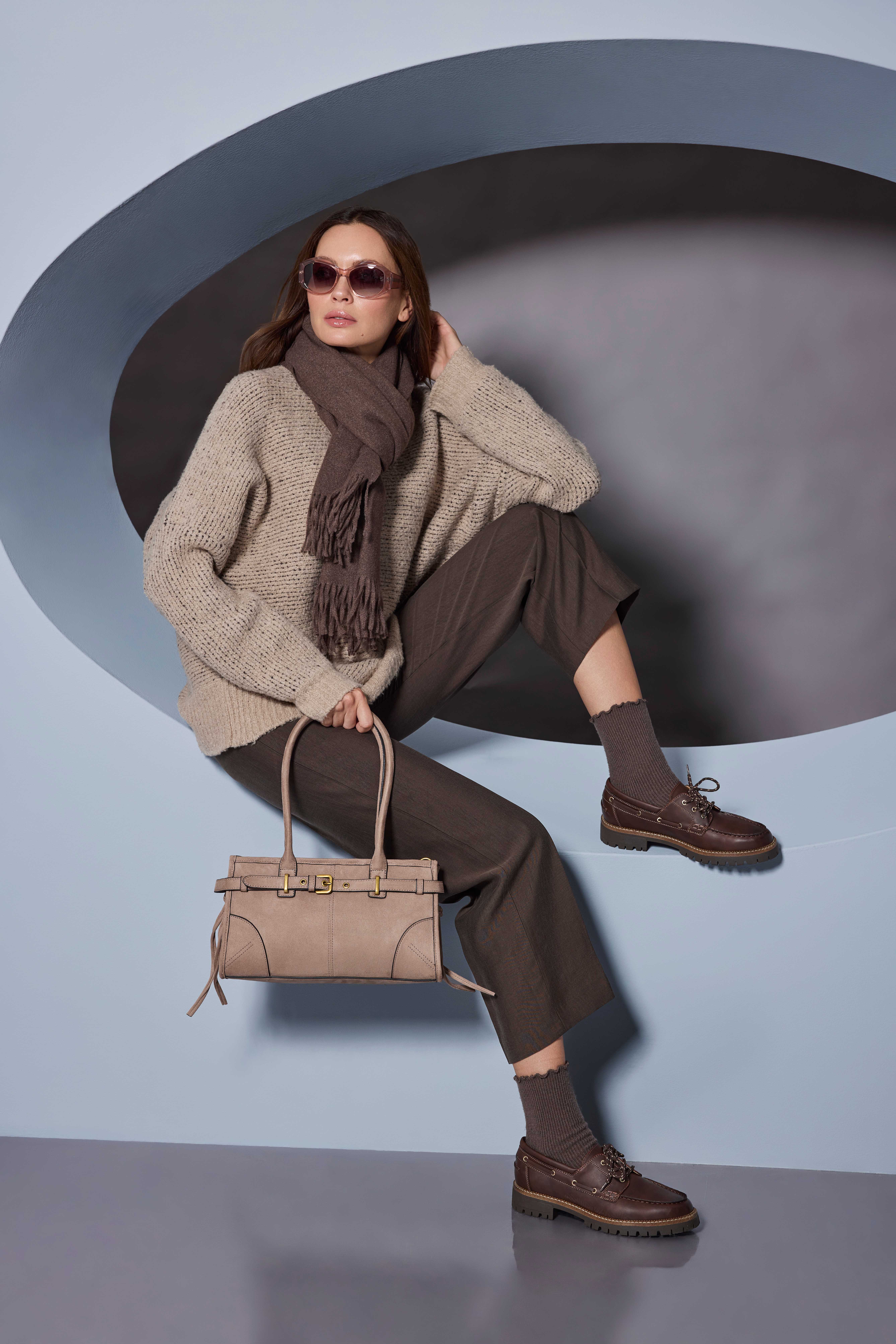 Frau mit Sonnenbrille, Strickpullover und Schal sitzt mit Handtasche vor grauem Hintergrund