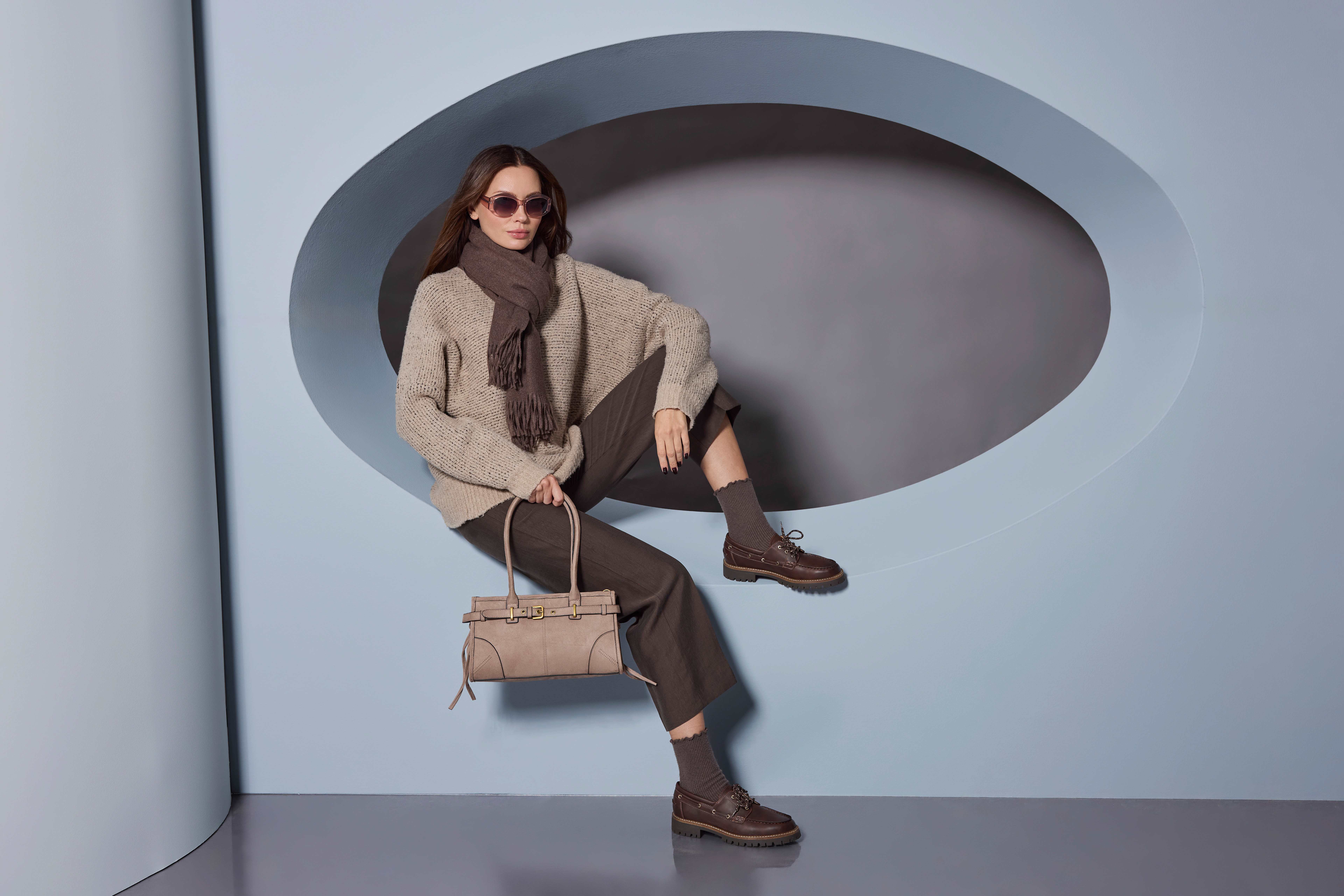 Eine Frau mit Sonnenbrille sitzt in einer Wandnische und trägt einen beigen Pullover, Schal und braune Kleidung.
