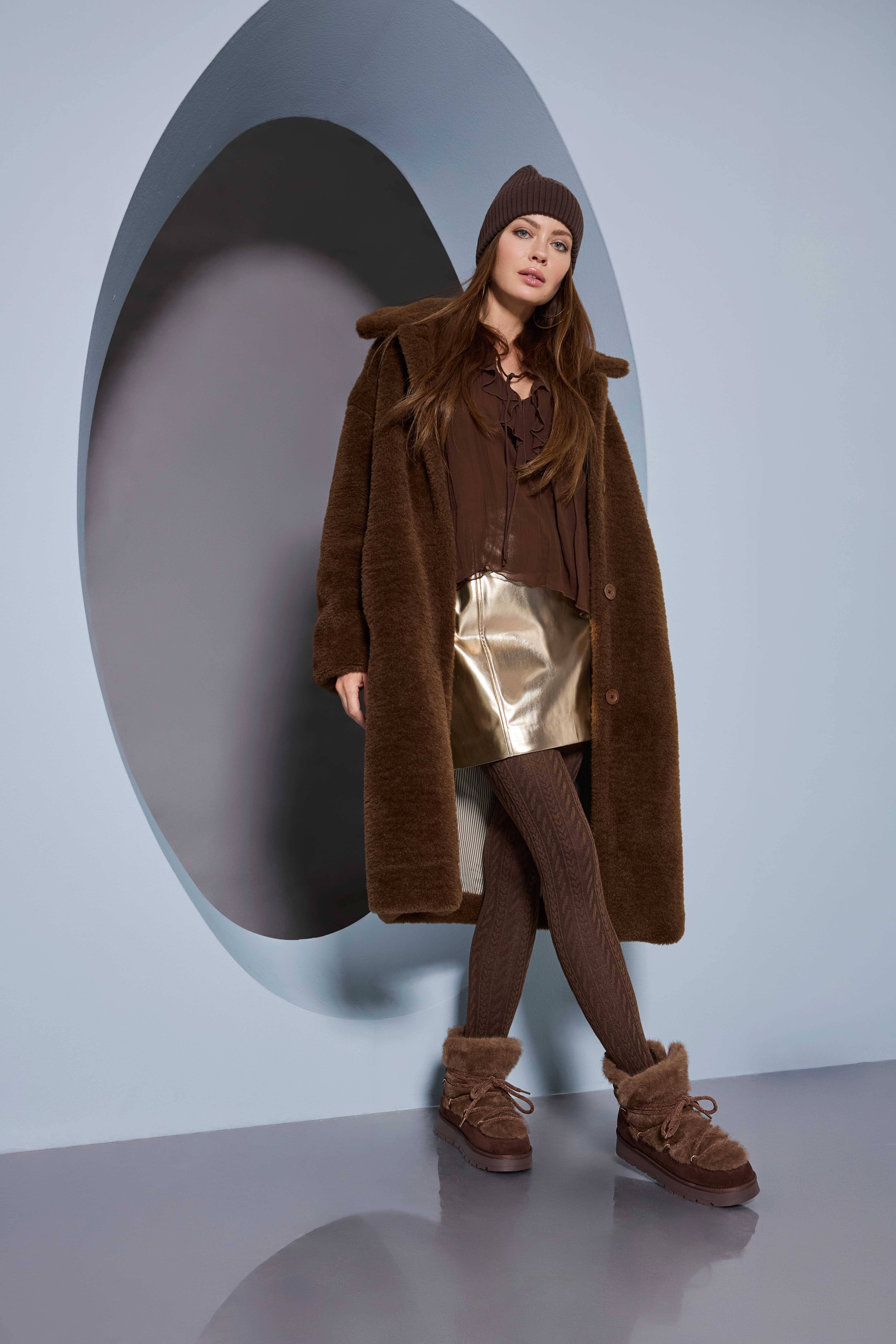 Frau in braunem Wintermantel, Strickmütze, Metallic-Rock, Strumpfhose und gefütterten Boots vor einer modernen Wand