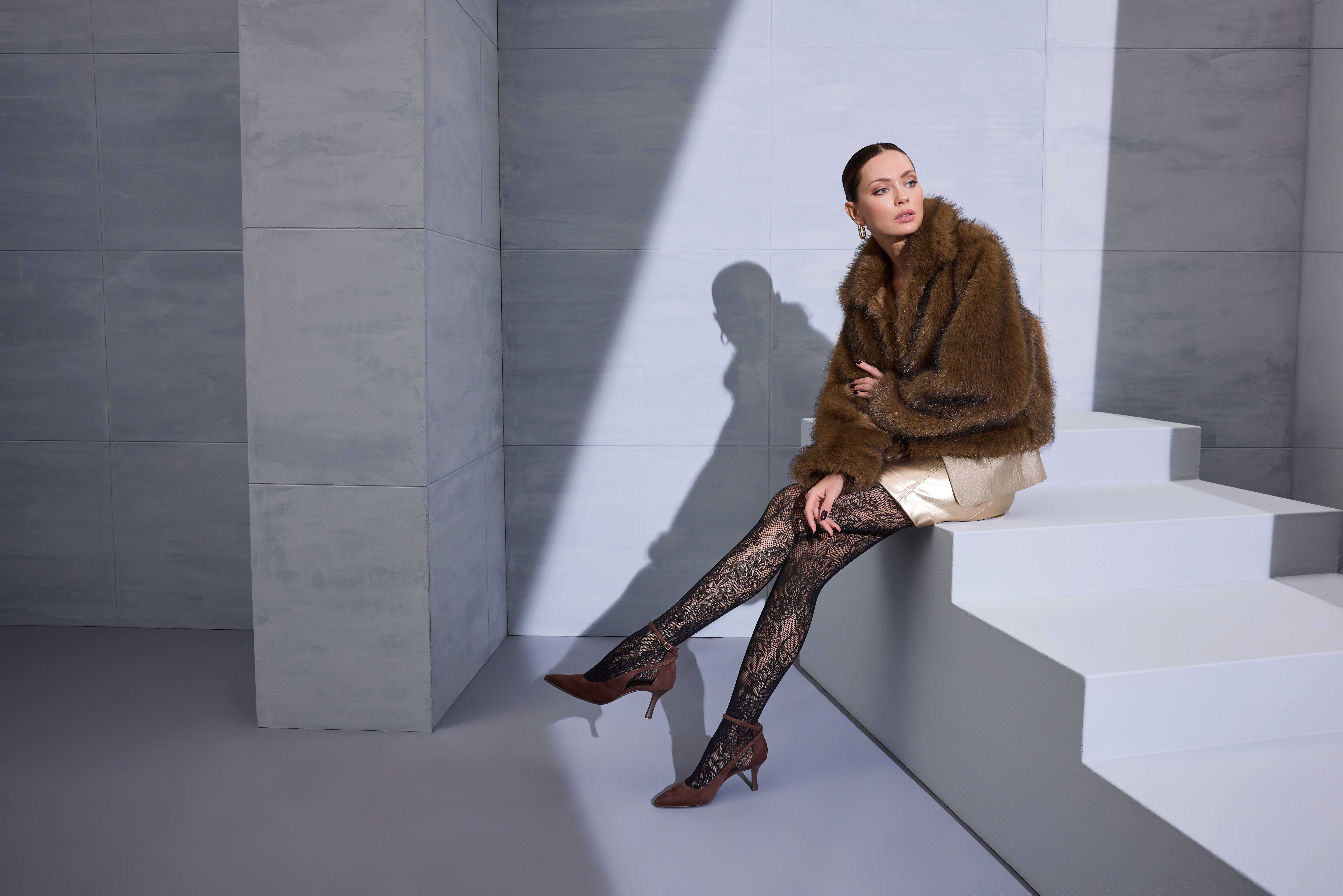 Eine Frau im Pelzmantel sitzt auf einer weißen Treppe und trägt Spitzenstrumpfhosen und braune Pumps.