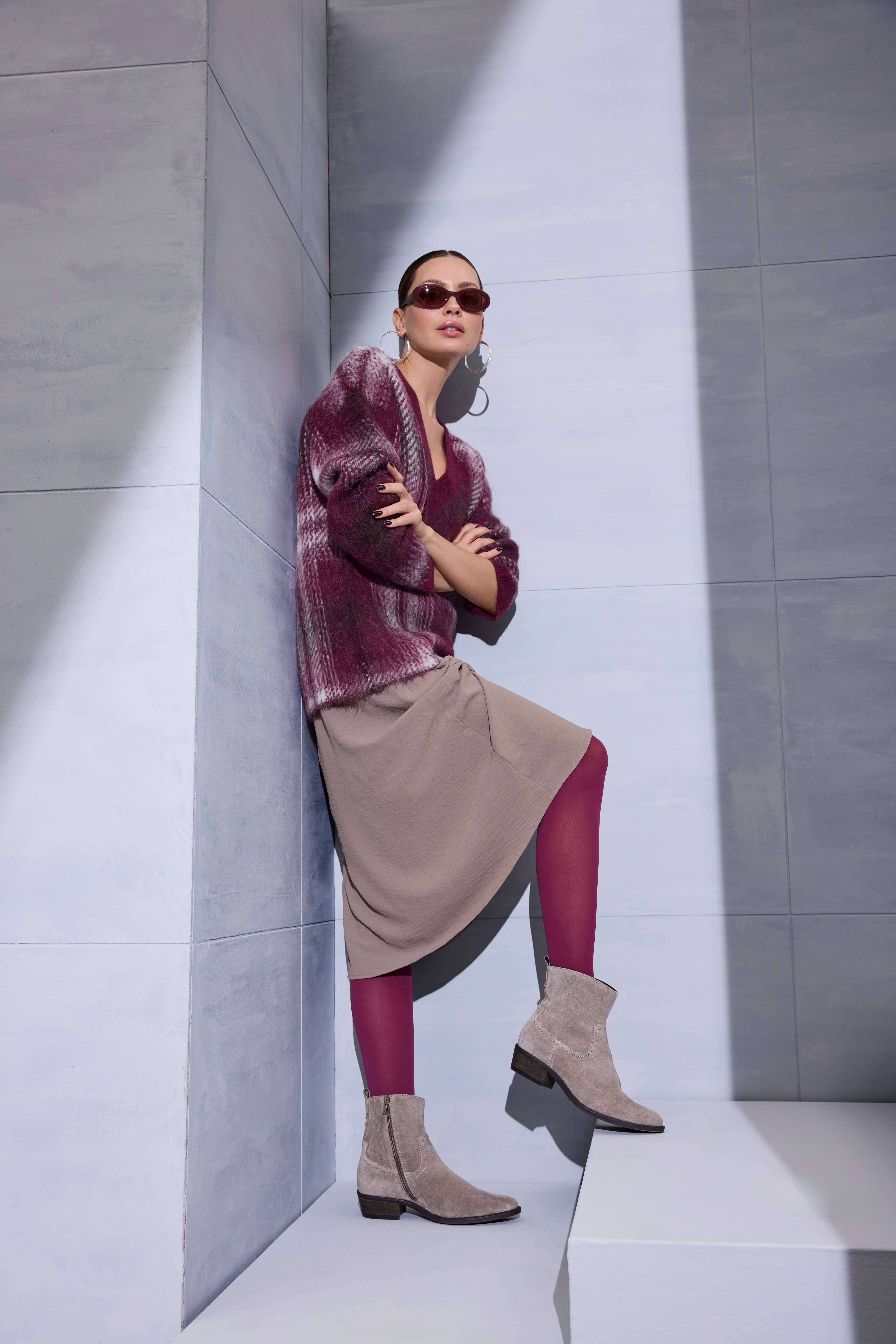 Eine Frau steht an einer modernen Betonwand und trägt eine burgunderrote Strickjacke, einen beigefarbenen Rock, farblich passende Strumpfhosen und beige Stiefeletten.