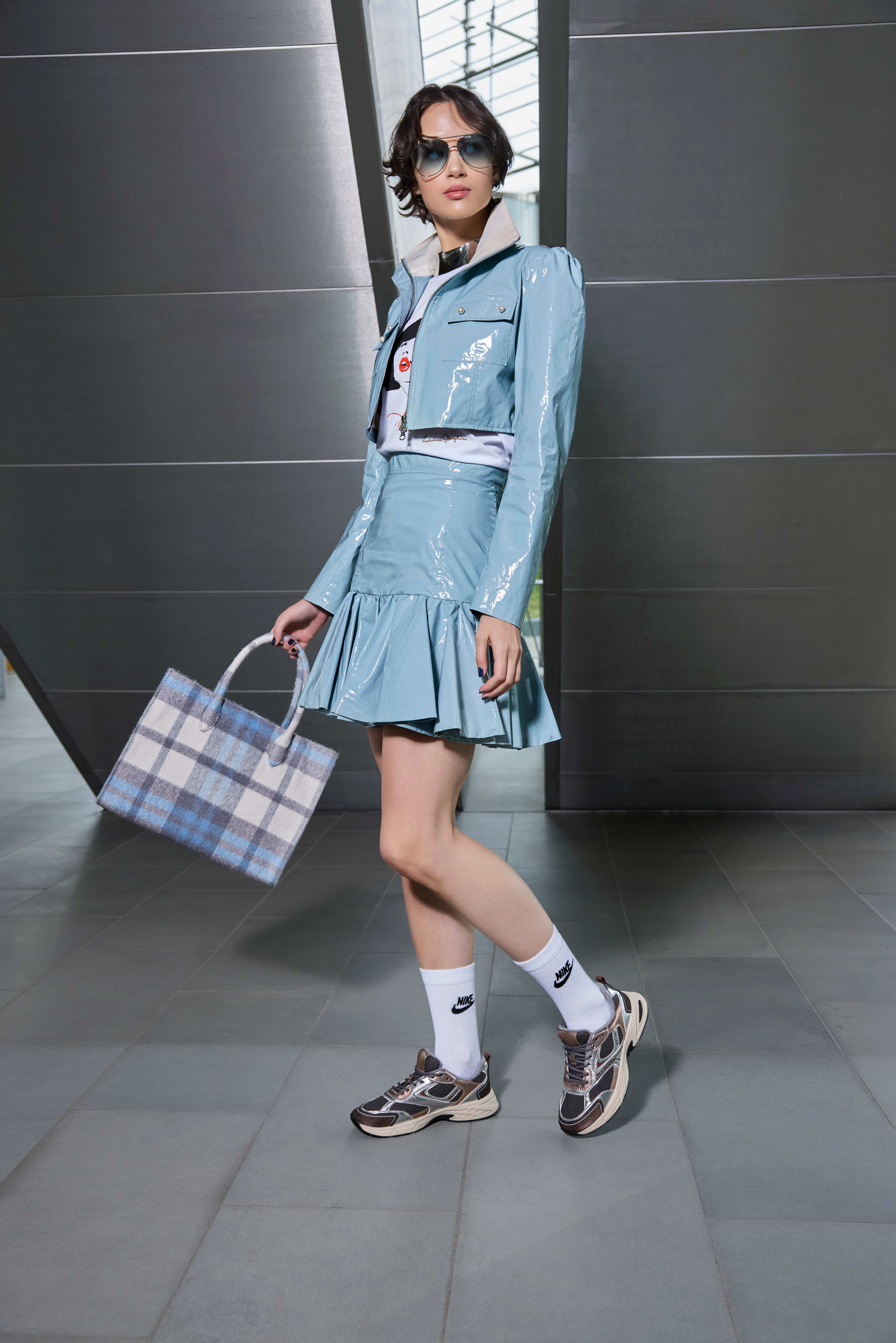 Frau im hellblauen Lack-Set mit Volantrock trägt Metallic-Sneaker, weiße Tennissocken und Karo-Shopper