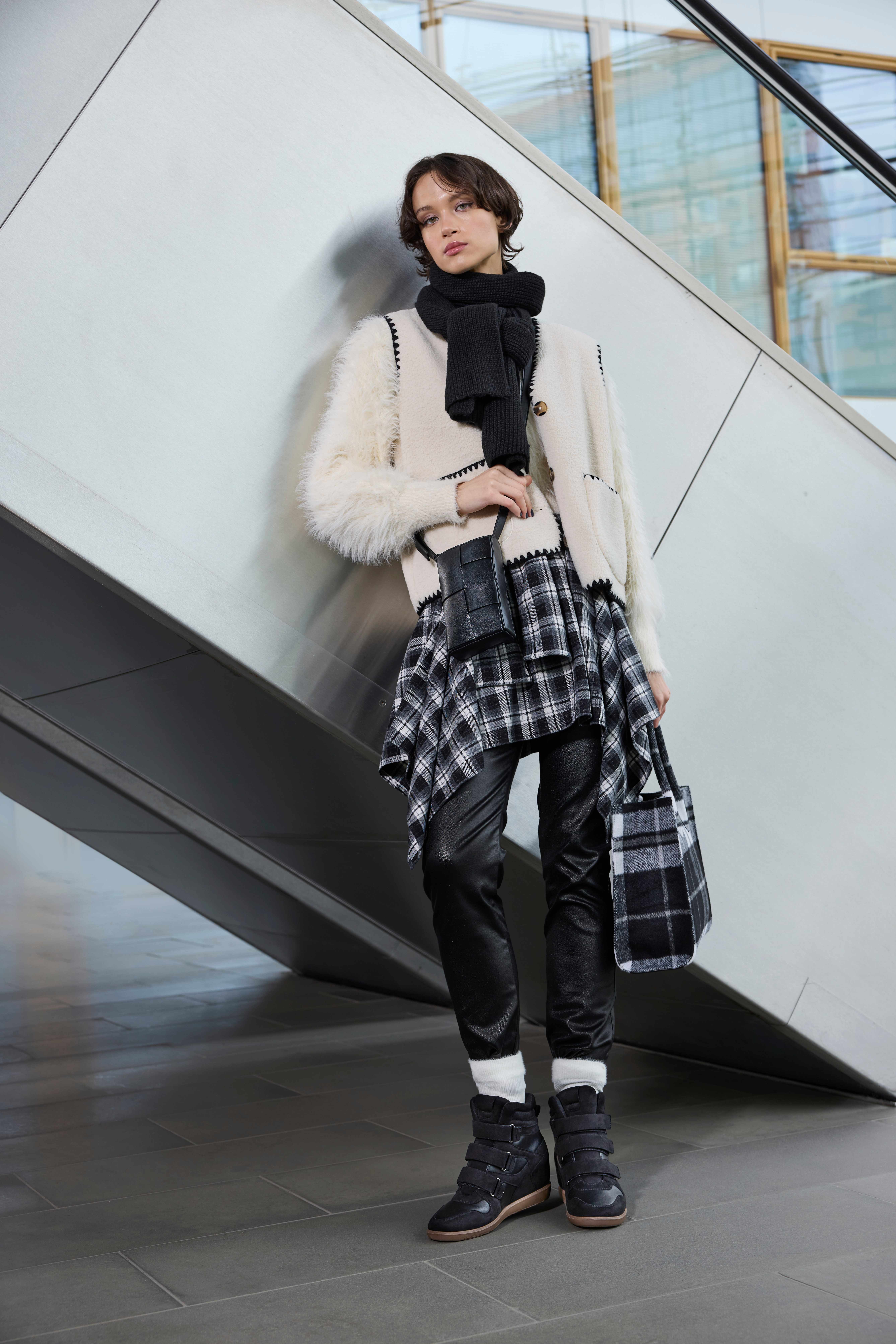 Eine Person in schwarz-weißer Kleidung mit kariertem Rock und schwarzen Sneaker-Wedges steht vor einer modernen Rolltreppe.