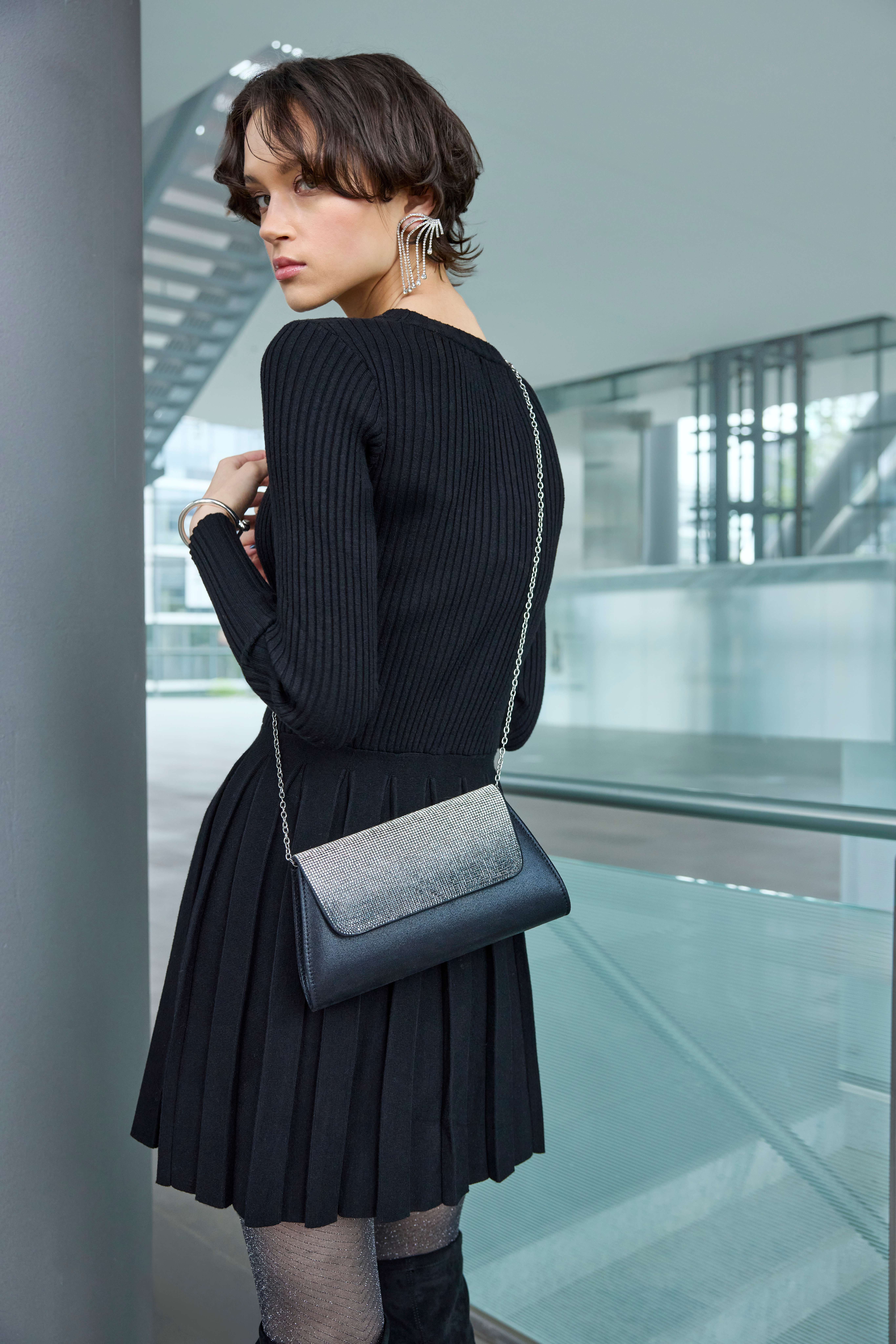 Frau in schwarzem Rippstrickkleid mit Plisseerock trägt Clutch mit Strasssteinen und Glitzerstrumpfhose