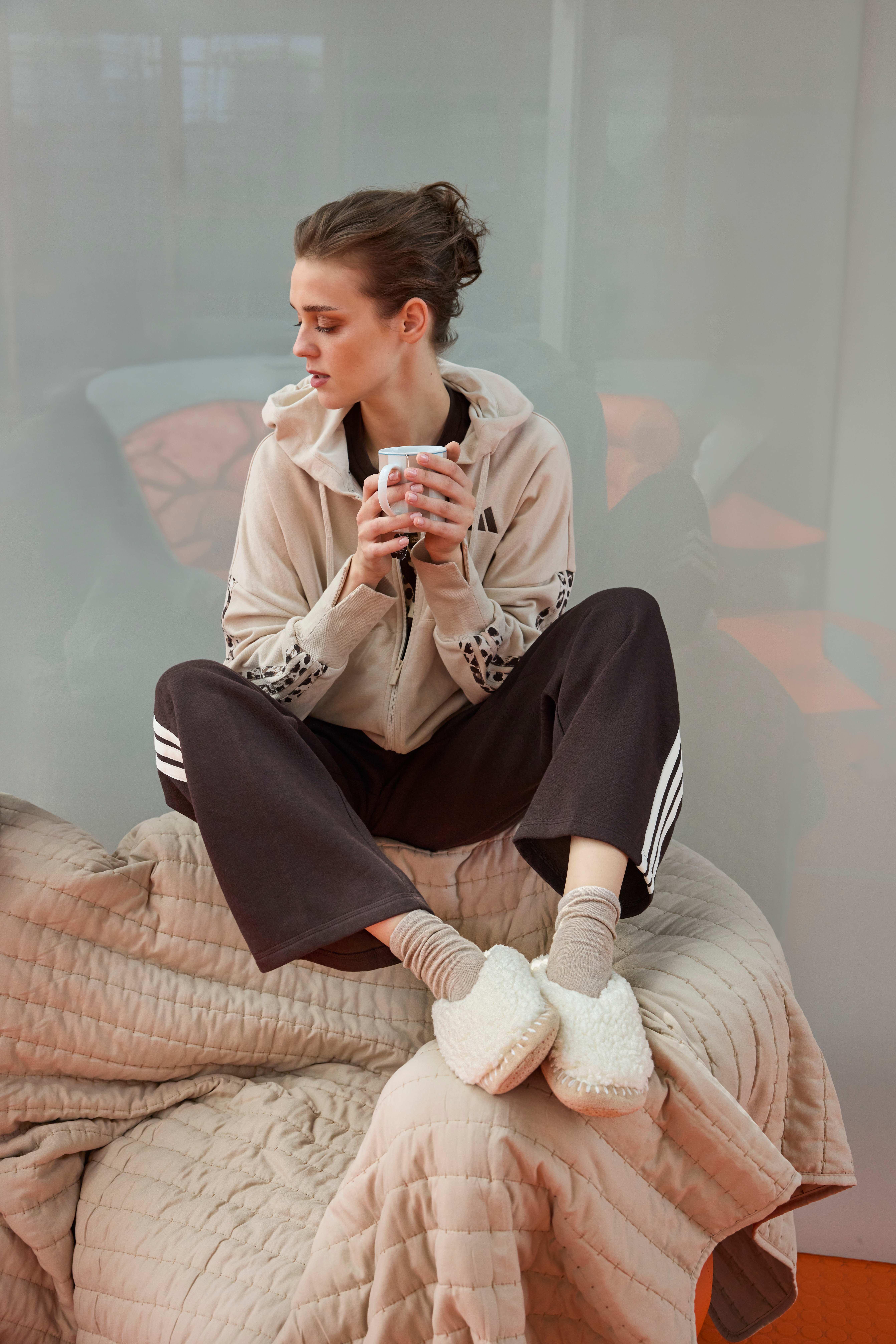 Frau sitzt auf Couch mit Tasse in Hand, trägt braune Hose, beige Zip-Jacke und cozy Boots