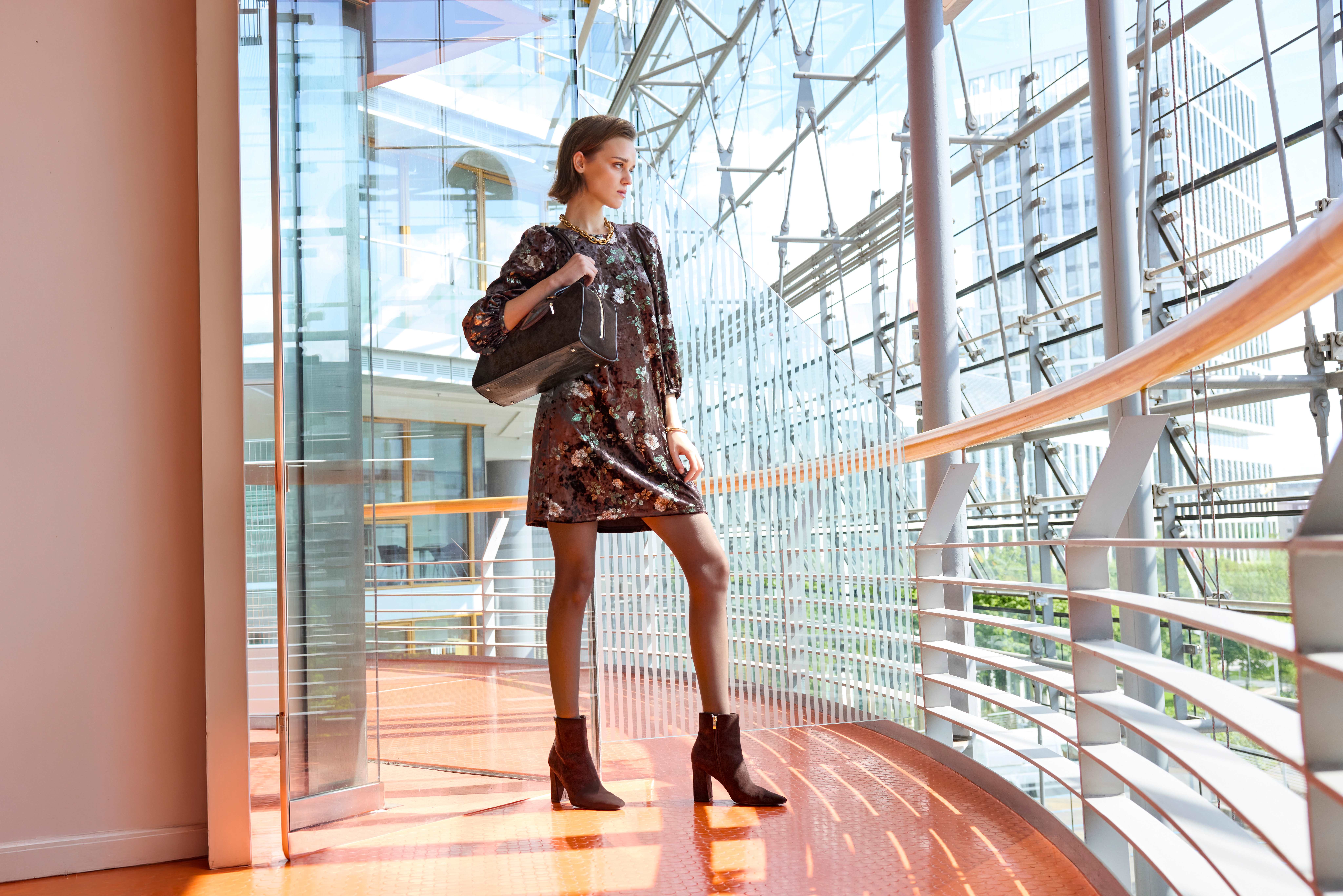 Frau im Samtkleid mit Blumenmuster, brauner Handtasche und Stiefeletten in Glasarchitektur