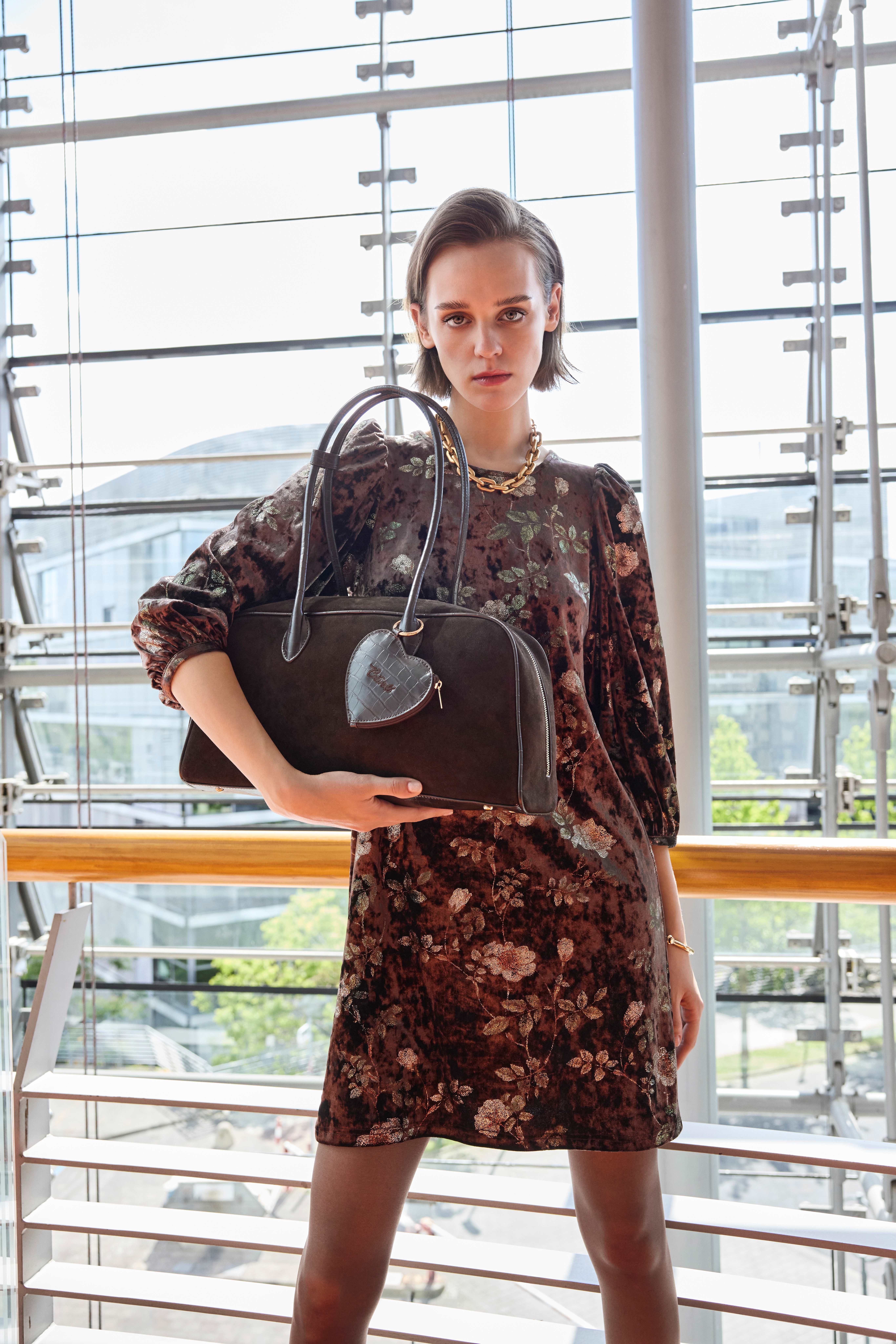 Model im Samtkleid mit Blumenmuster hält braune Bowling-Tasche mit Herzanhänger