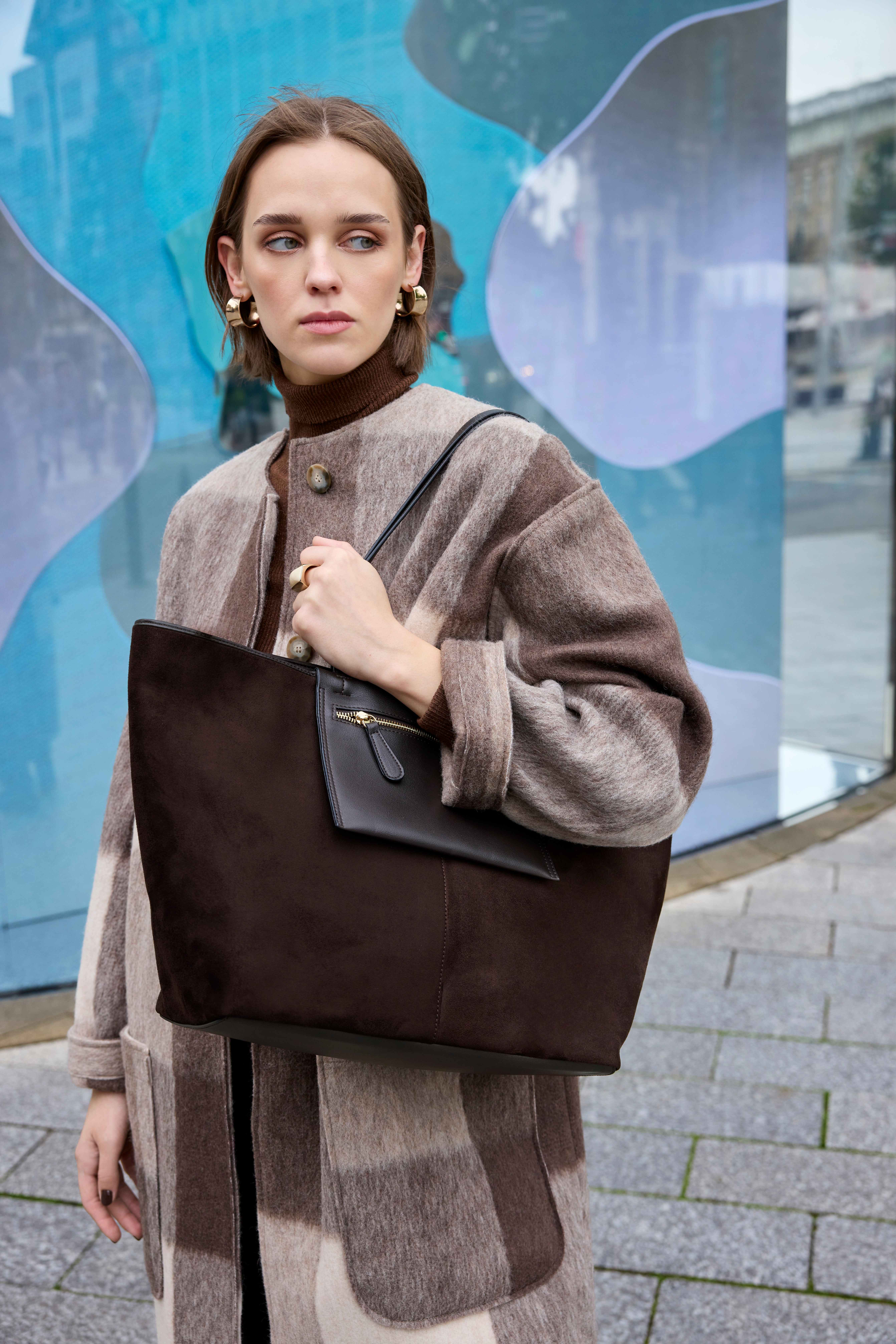 Frau im Wollmantel mit Karo-Muster trägt große Shopper-Tasche aus braunem Wildleder