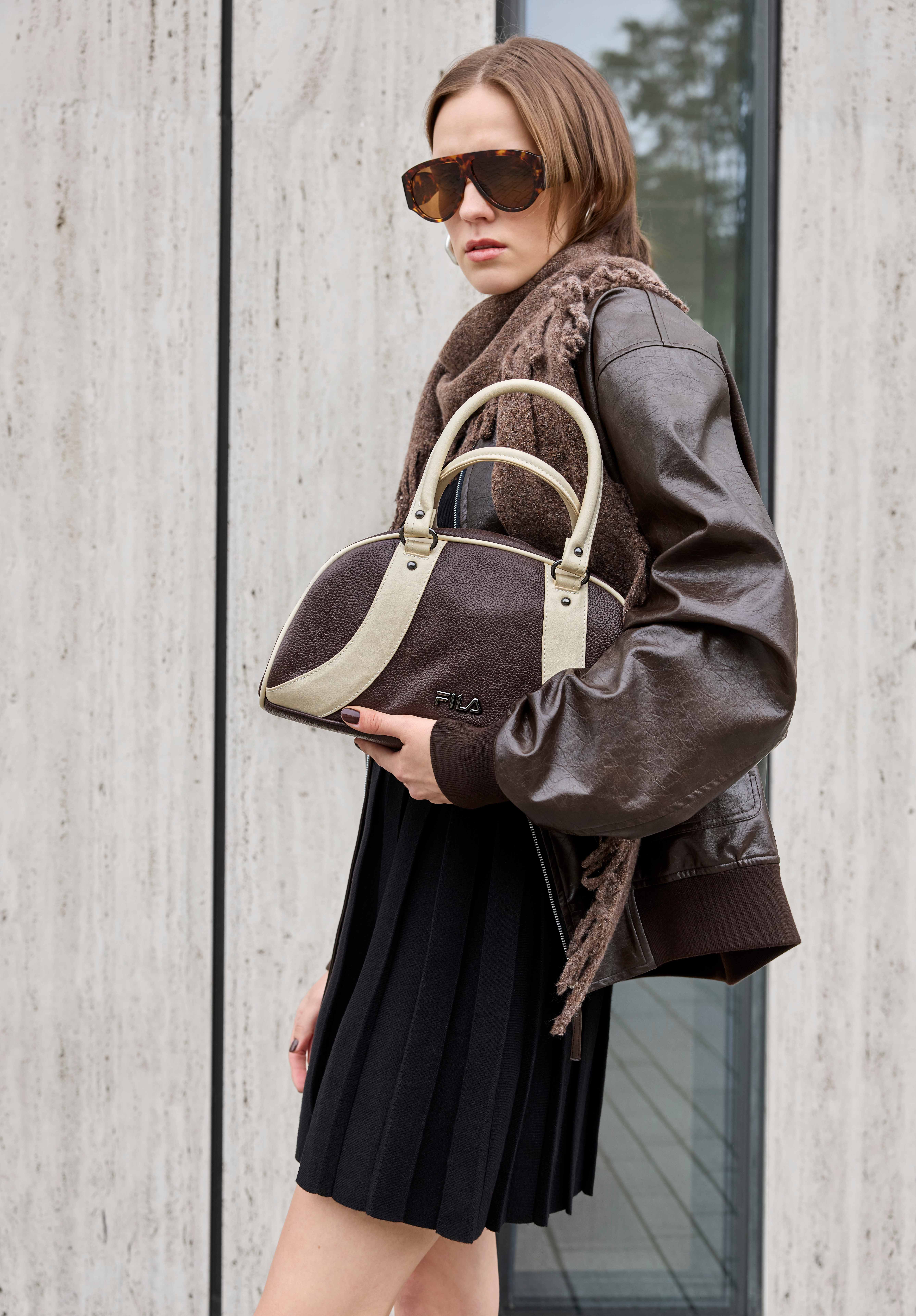 Model in brauner Lederjacke und Plisseerock trägt braune Fila-Bowlingtasche