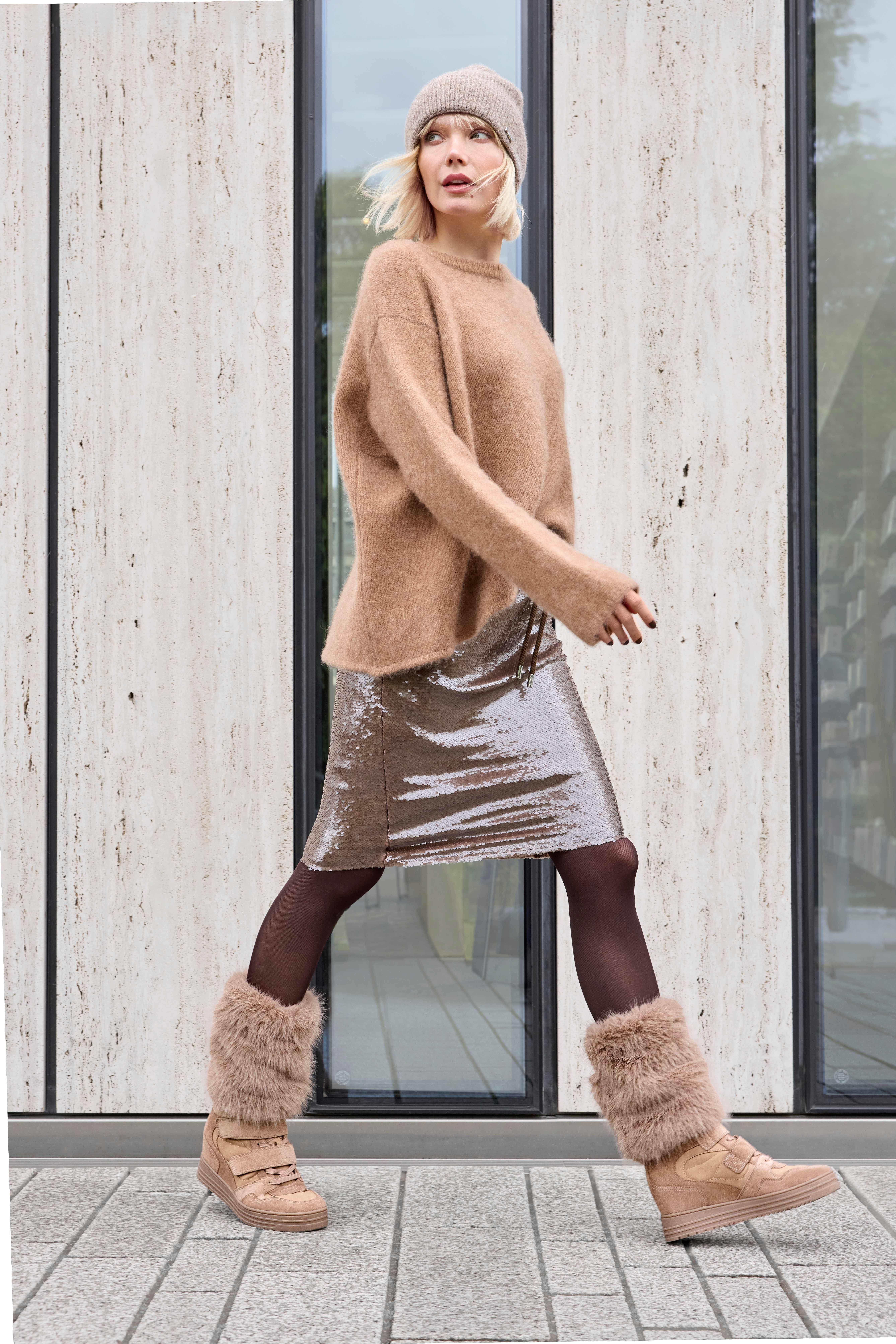 Eine Frau trägt einen beigefarbenen Strickpullover, einen glänzenden Rock und gefütterte Sneaker-Wedges mit Fellbesatz.