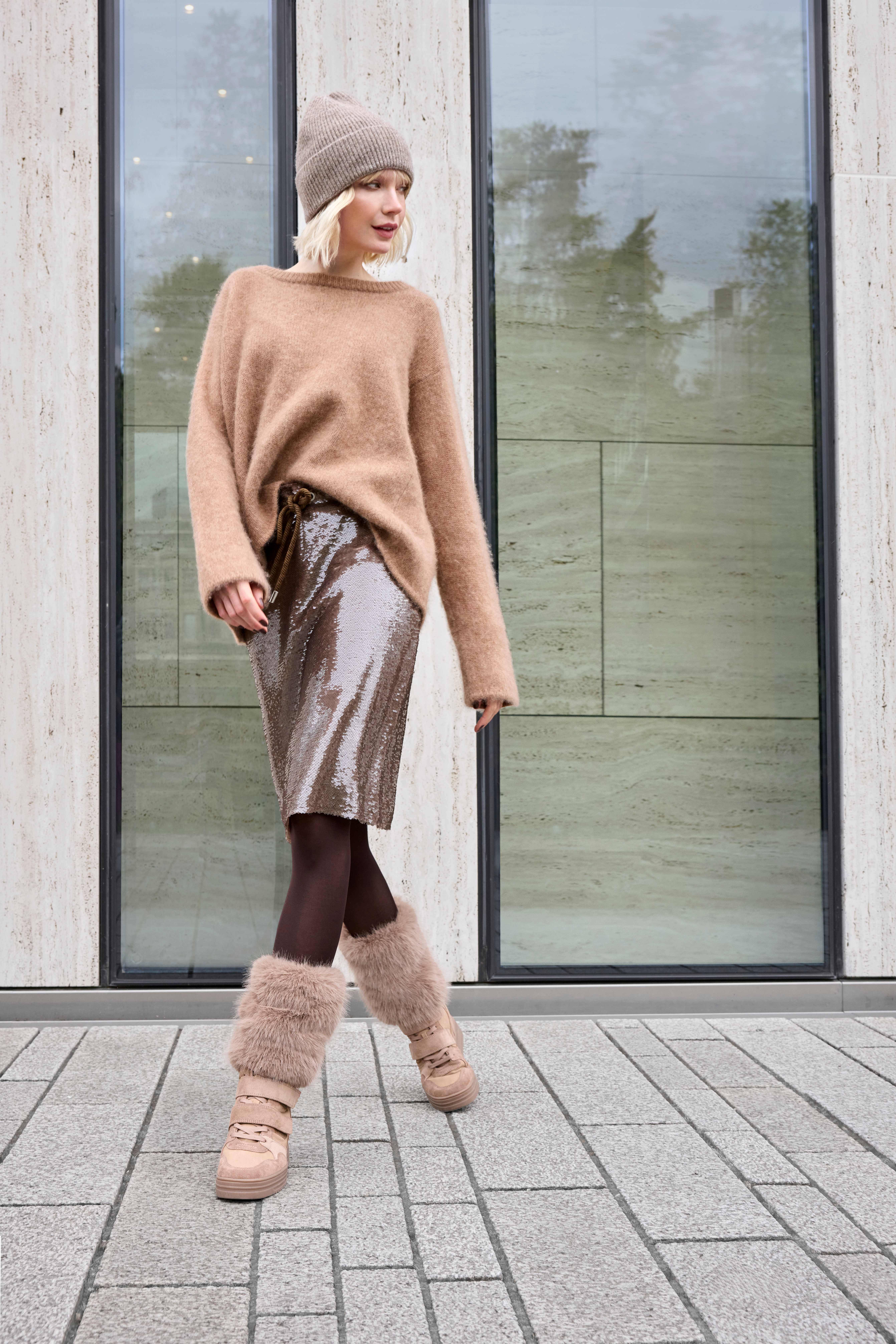 Frau trägt einen beigen Pullover, eine Strickmütze, einen glänzenden Rock, dunkle Strumpfhosen und Fellstulpen