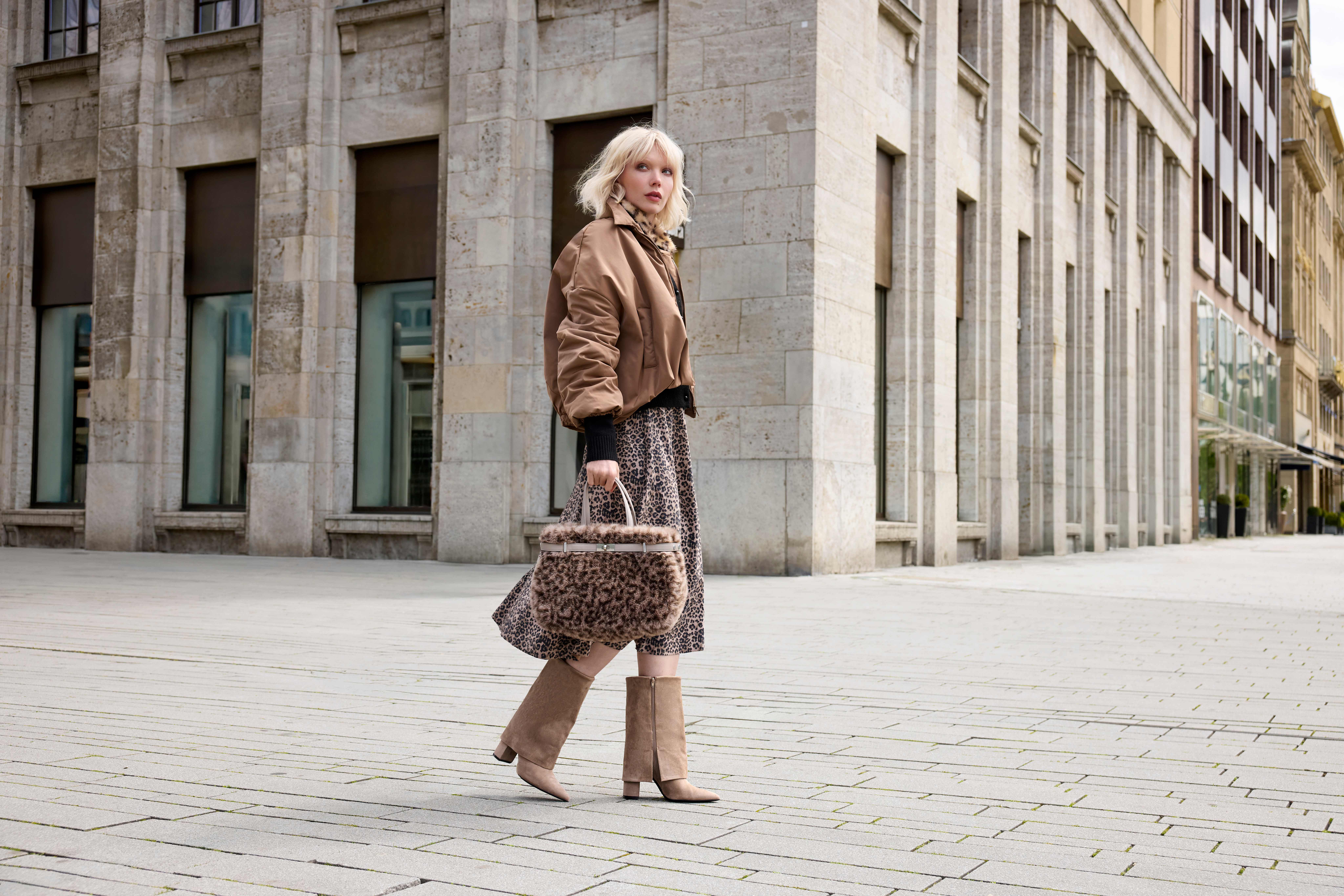 Eine Frau in gemusterter Kleidung mit brauner Jacke, Stiefeln und einer Fellhandtasche vor einem Stadthaus