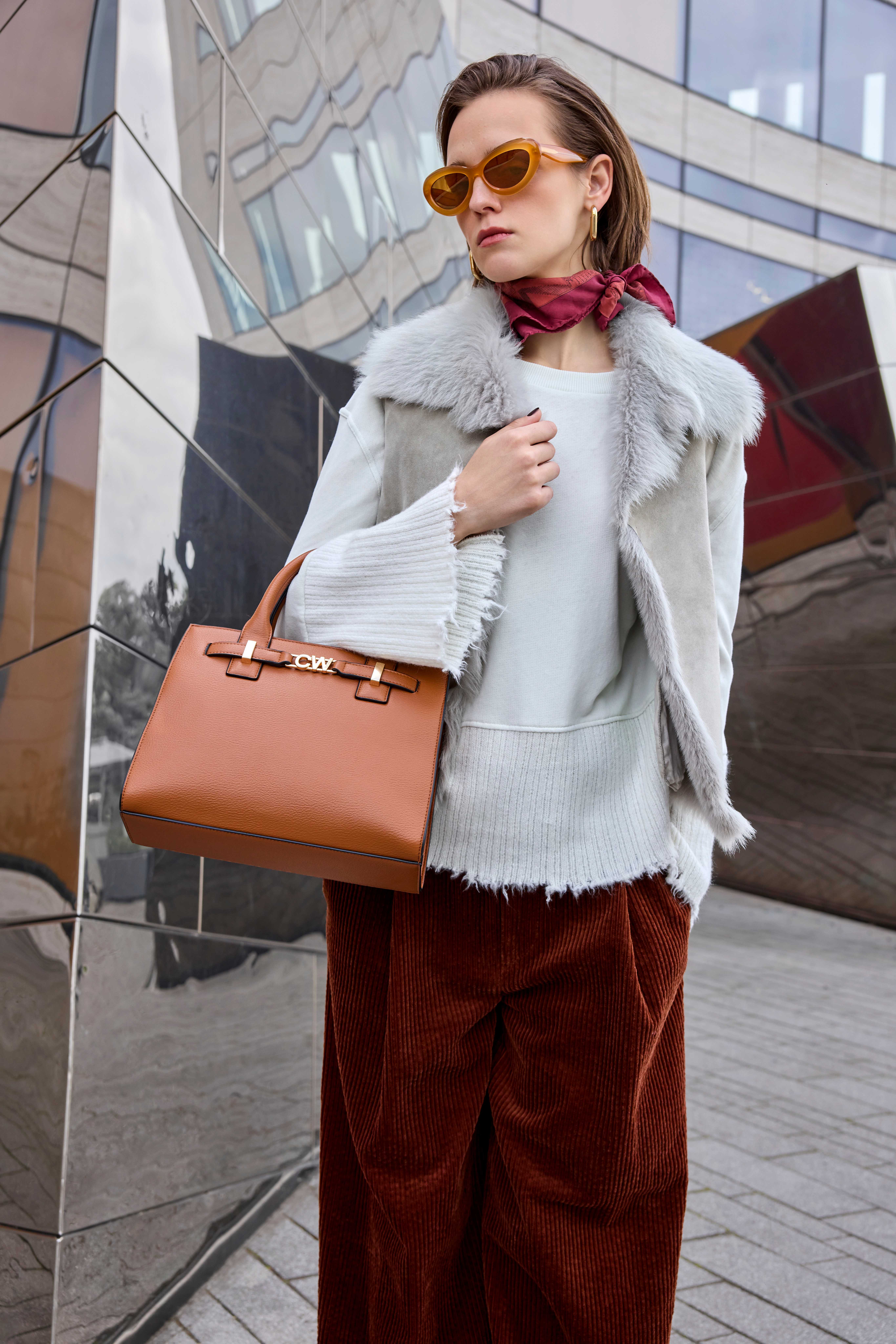 Eine Frau, die eine Sonnenbrille, eine mit Pelz besetzte Weste und eine braune Hose trägt und eine hellbraune Handtasche bei sich hat.