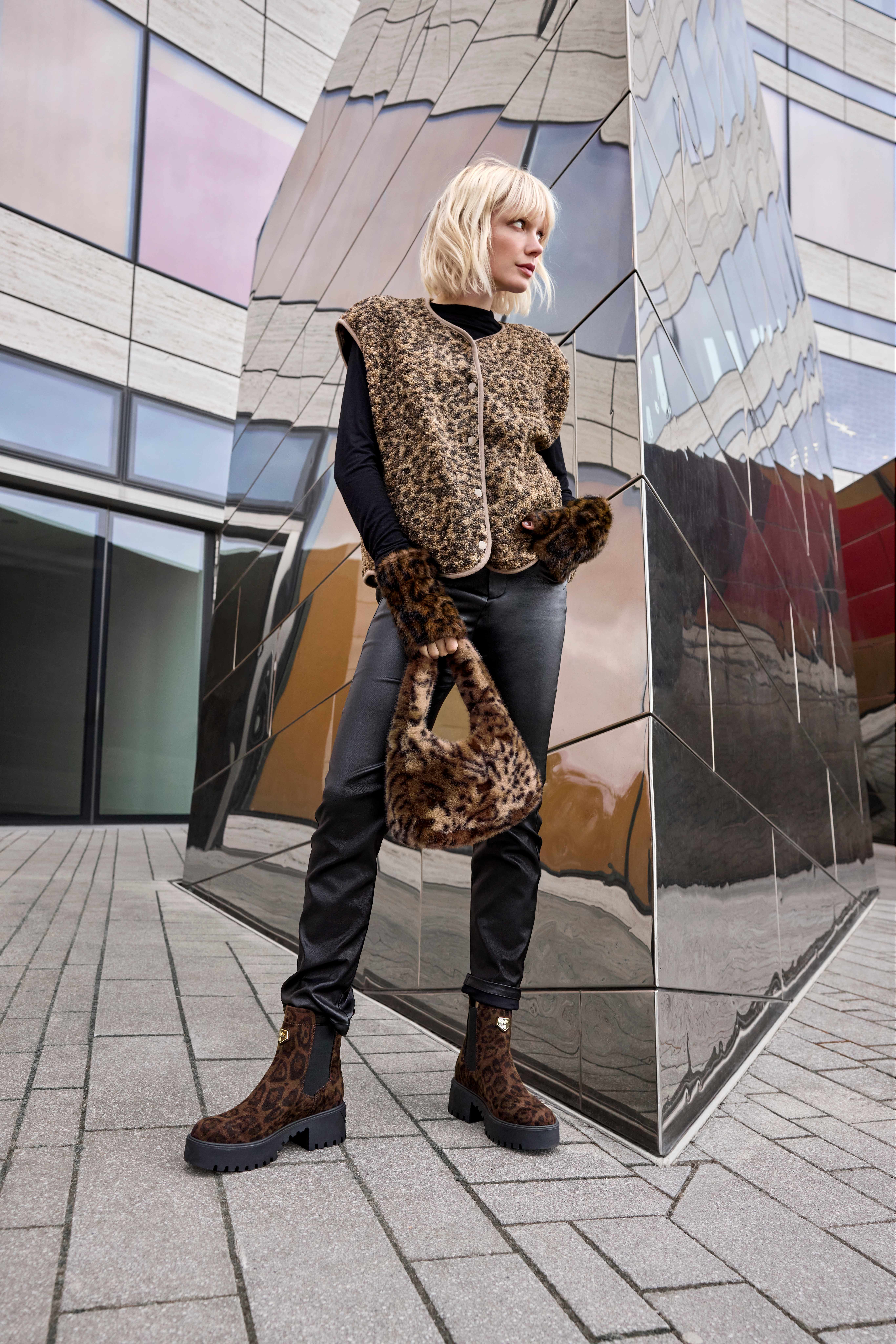 Eine Frau in Leopardenmuster-Weste, schwarzer Kleidung und Stiefeletten vor einem modernen Gebäude