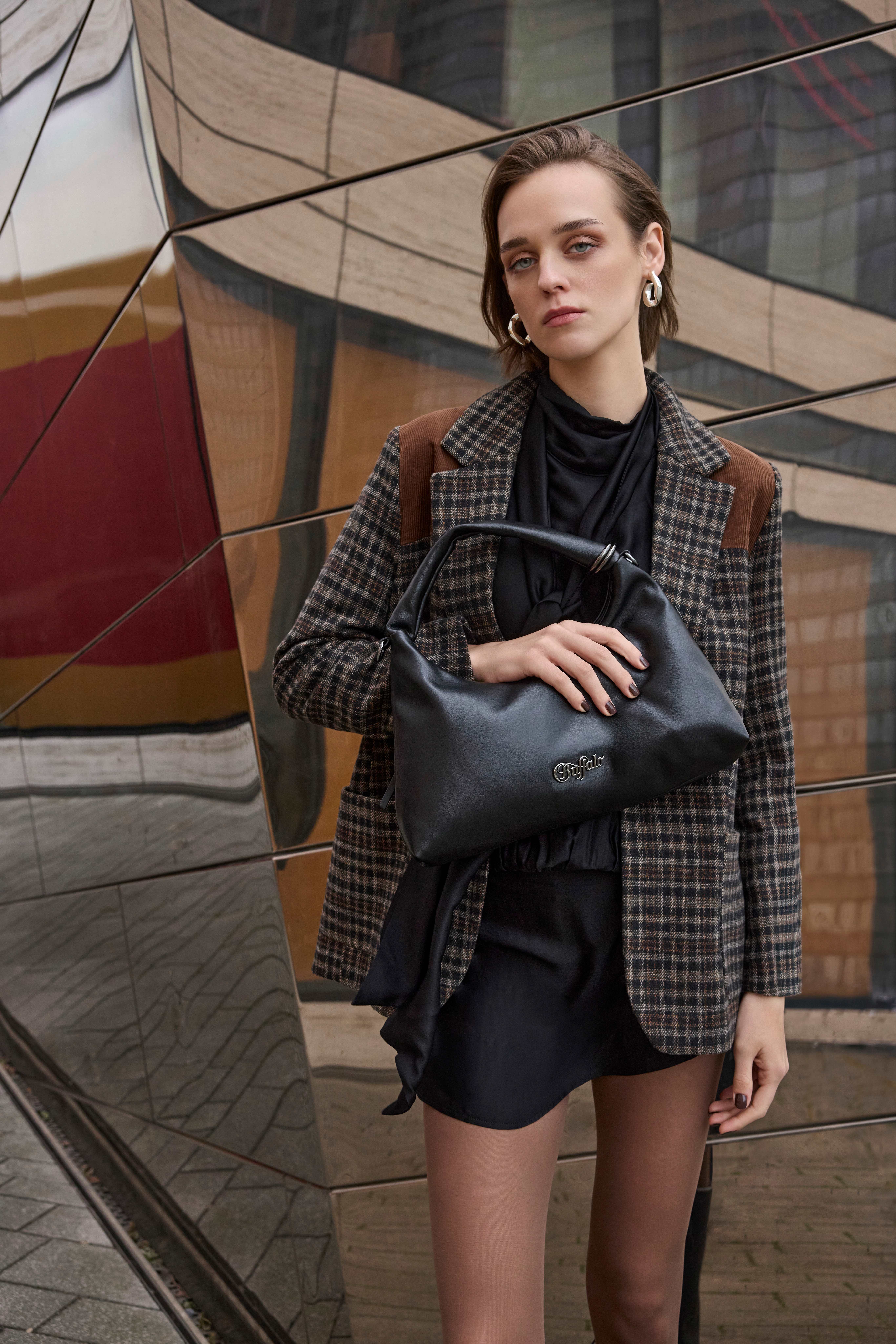 Frau in kariertem Blazer, schwarzem Outfit und mit großer schwarzer Handtasche vor einer modernen Fassade