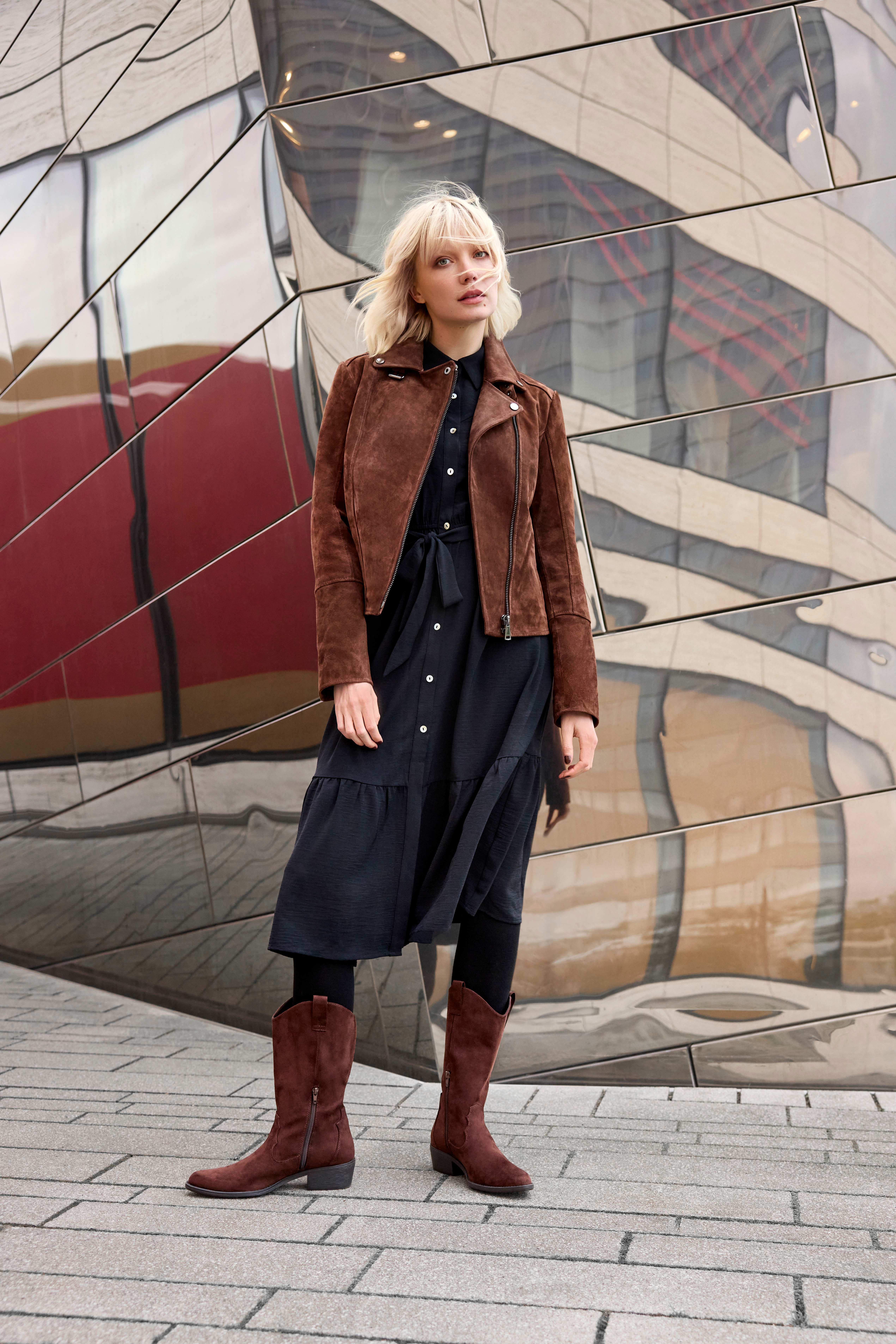 Eine Frau trägt hohe braune Western Boots zu einem schwarzen Kleid und einer braunen Lederjacke