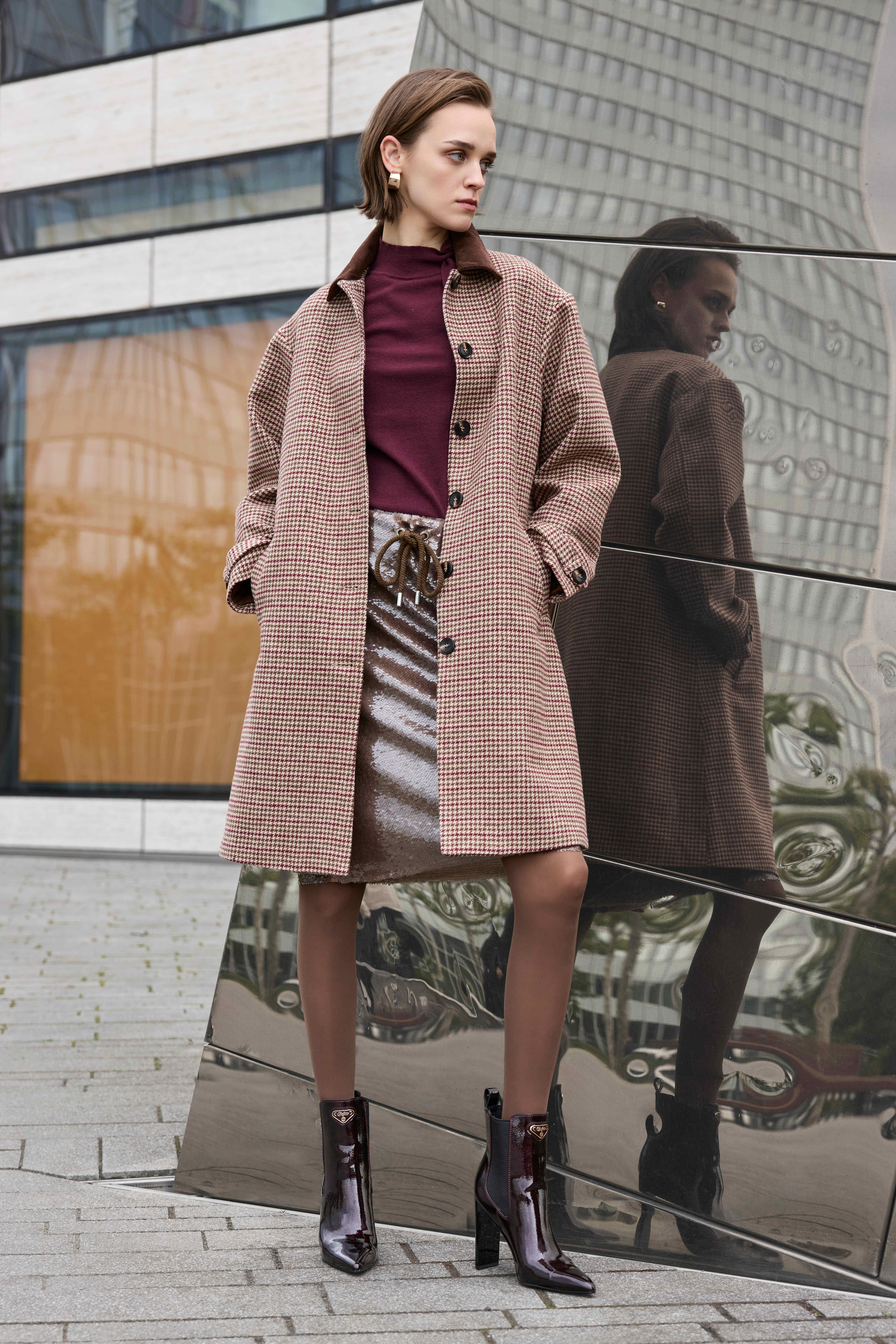 Eine Frau steht vor einem modernen Gebäude und trägt einen karierten Mantel, einen burgunderfarbenen Pullover, einen Rock und Stiefeletten.