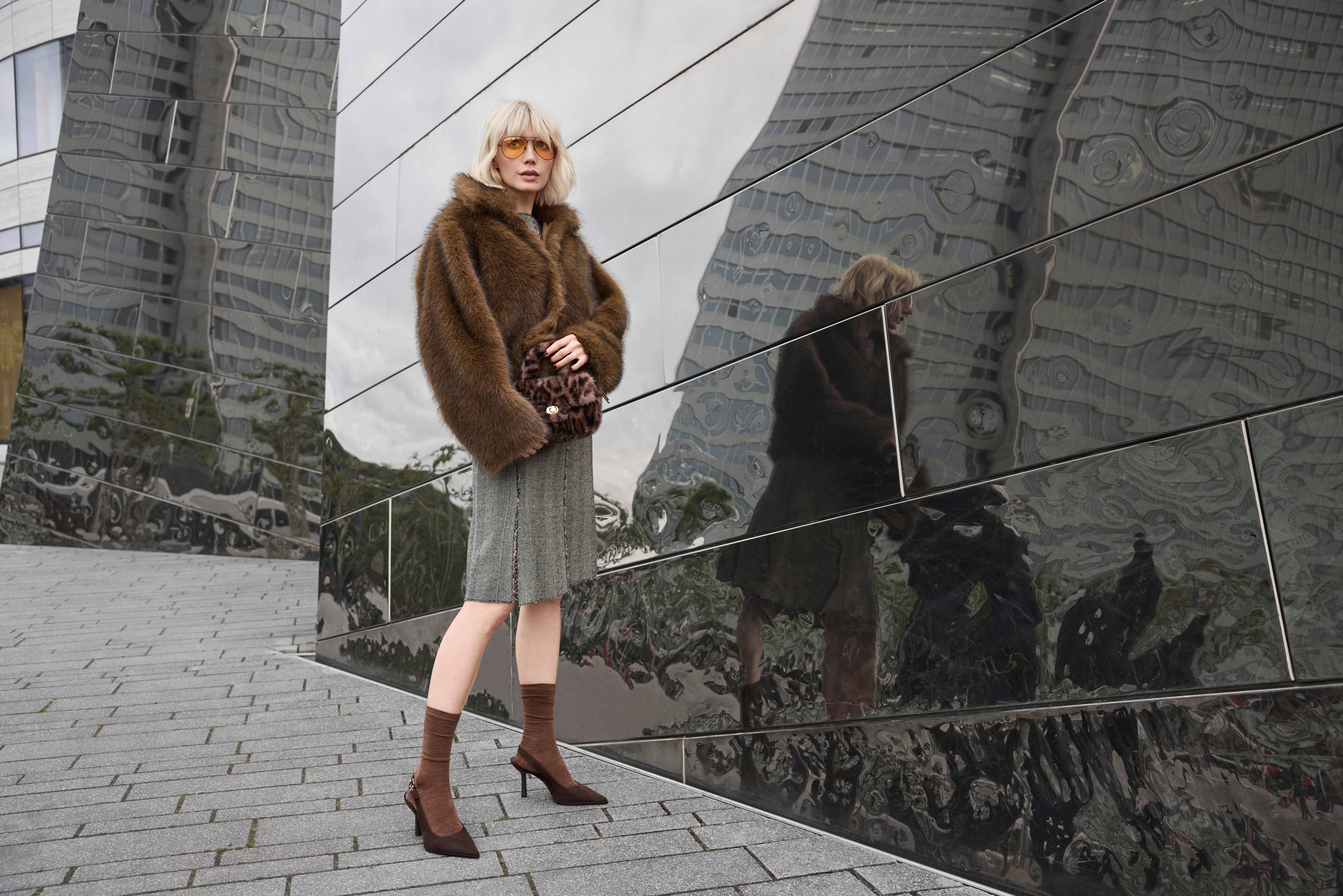 Frau trägt braune Felljacke im Boho-Style, mit Strickkleid und Slingback-Pumps vor Spiegelfassade