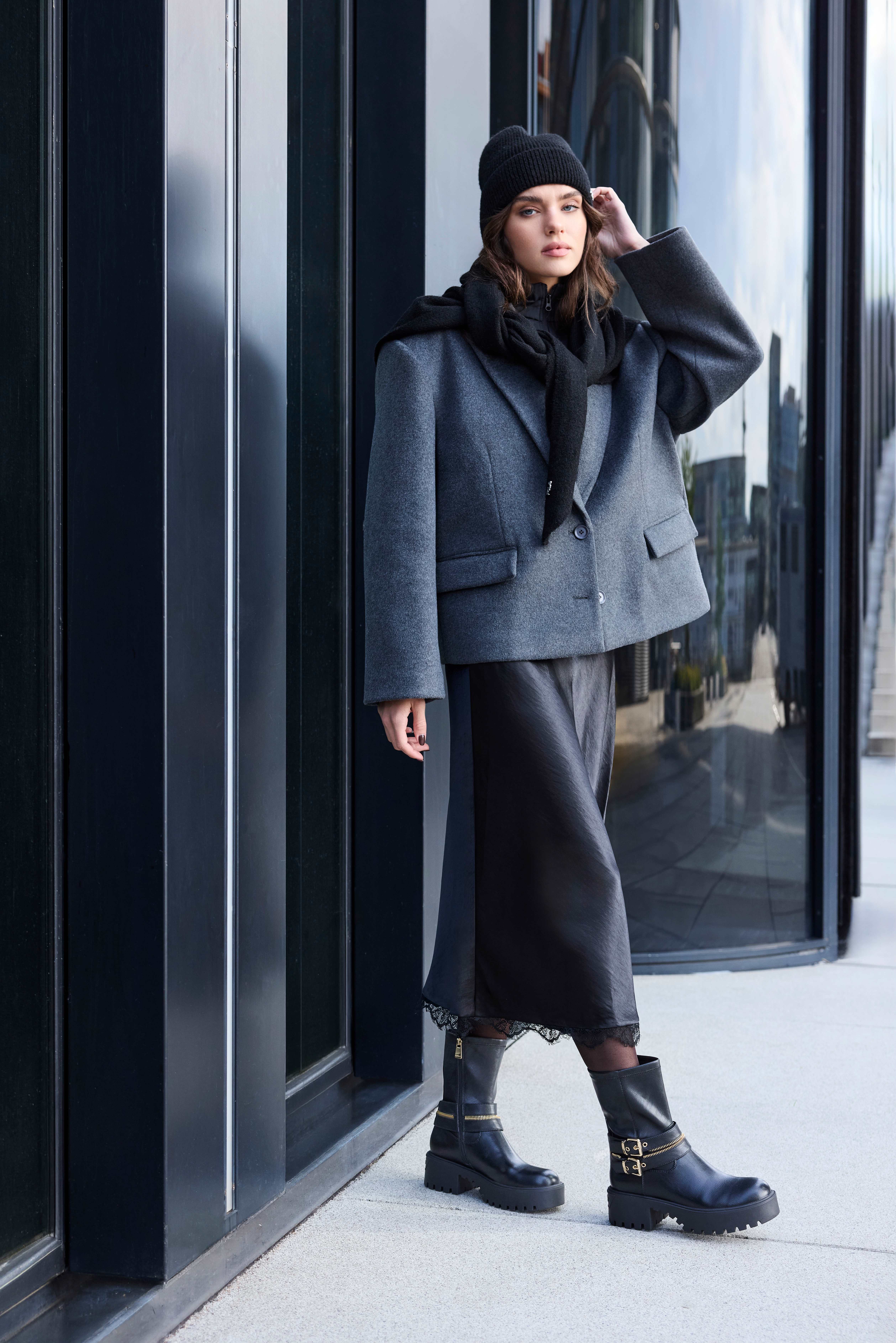 Frau in grauem Wollblazer, Beanie und schwarzem Satinrock trägt Biker Boots mit Schnallen und Profilsohle