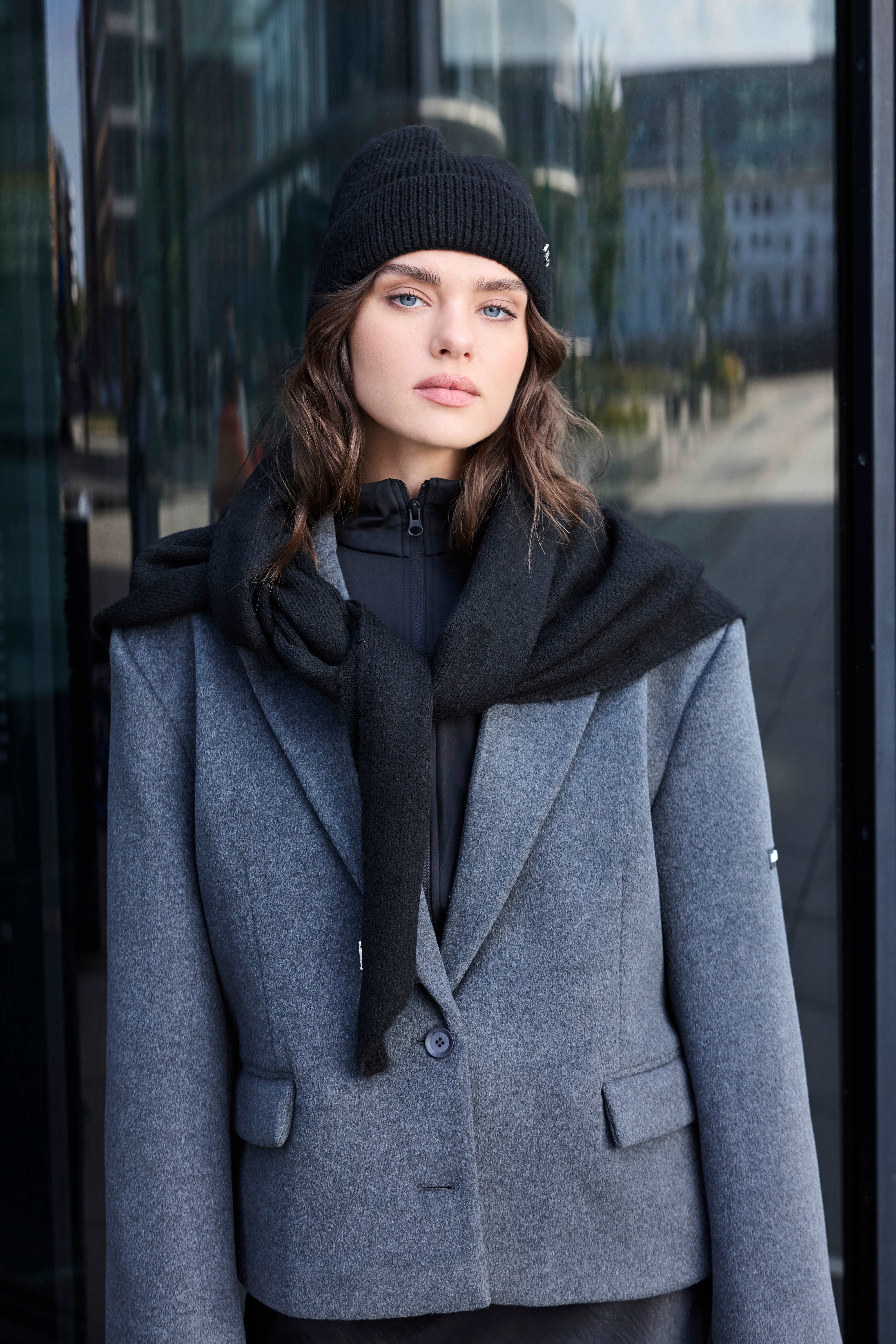 Frau steht in grau-schwarzem Layering-Outfit aus Blazer, Pullover und Beanie