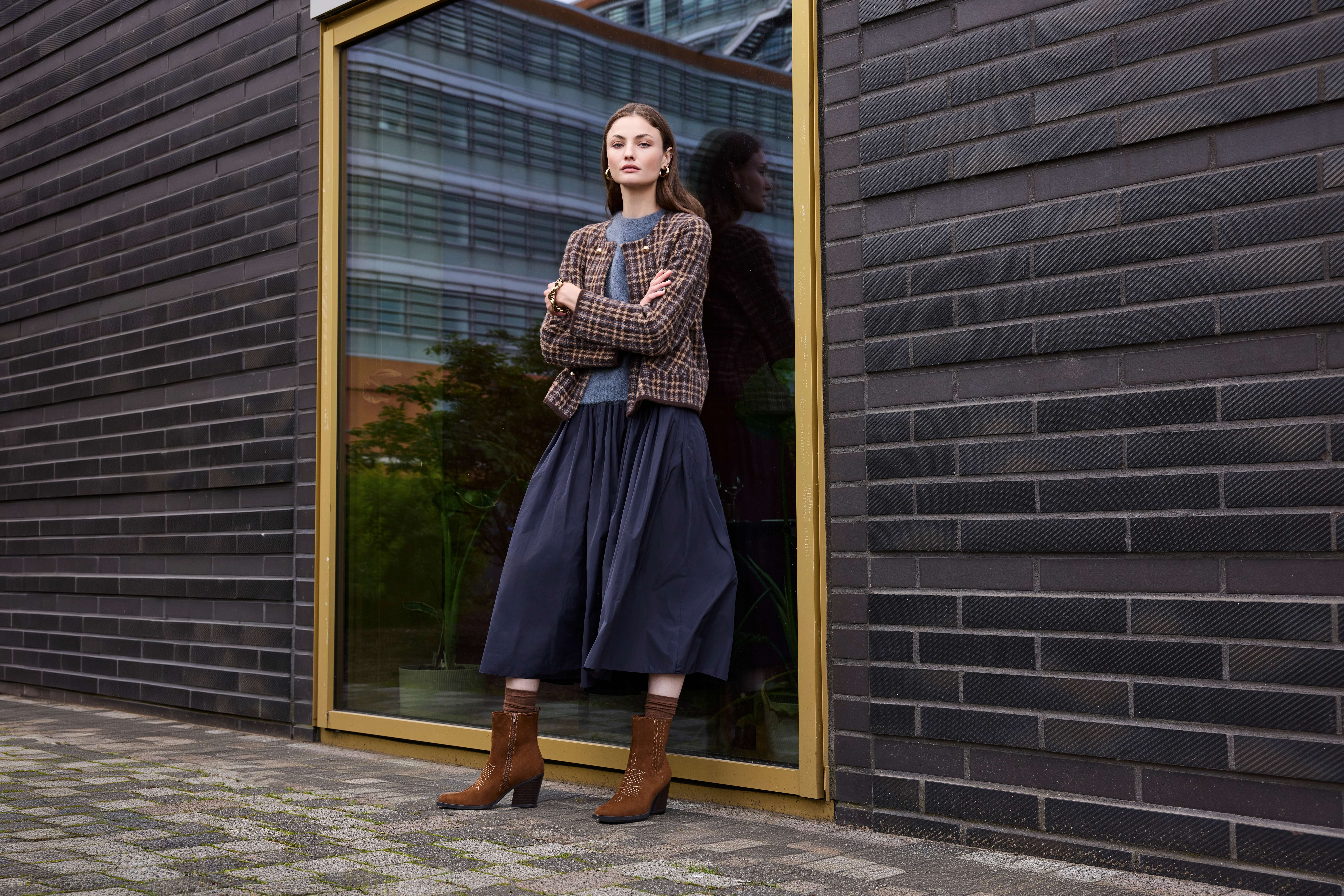 Eine Frau posiert in blauem Rock, braun karrierter Jacke und braunen Western Boots