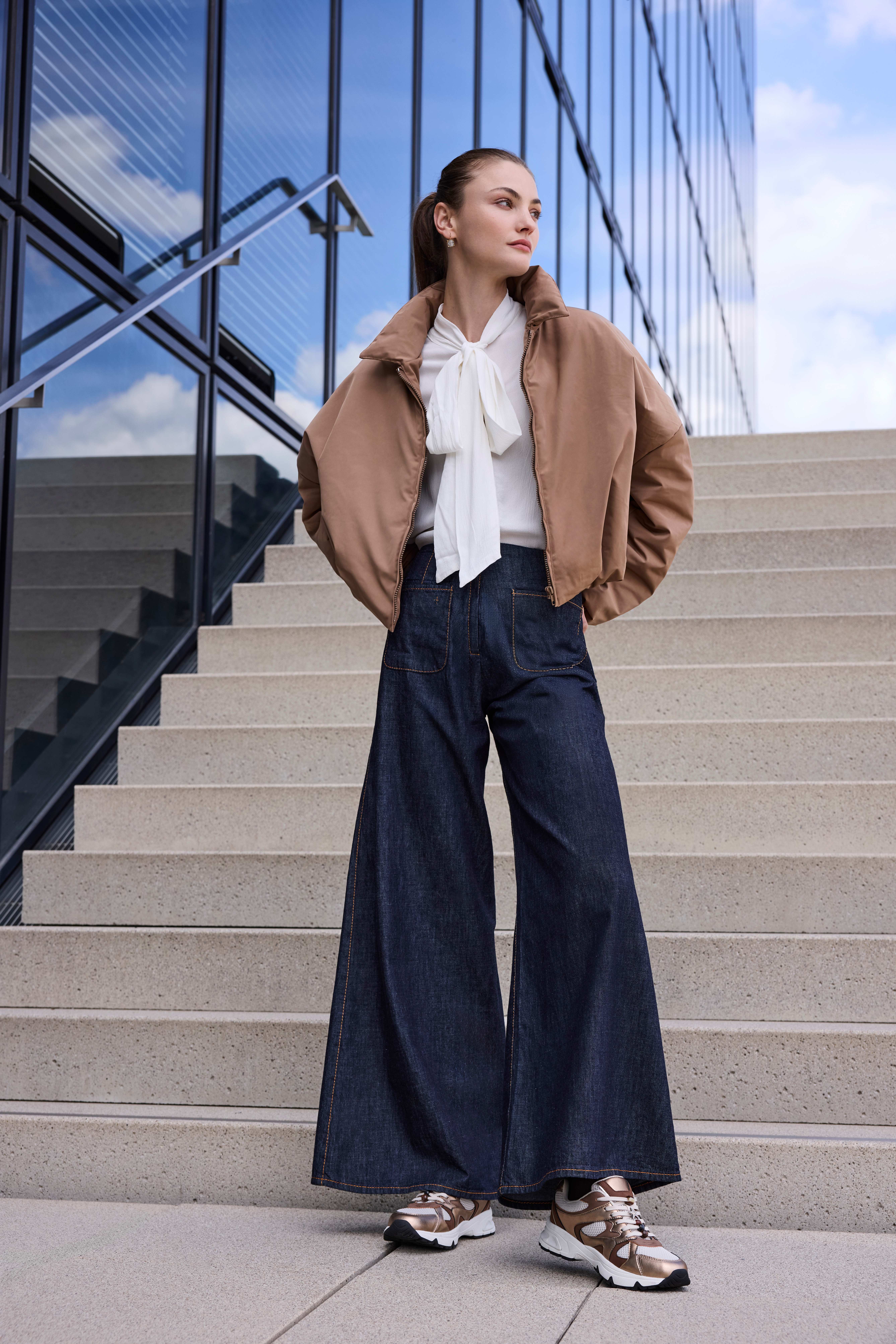 Frau in beigem Blouson, Schluppenbluse und Marlene-Jeans trägt Chunky Sneaker in Roségold-Metallic
