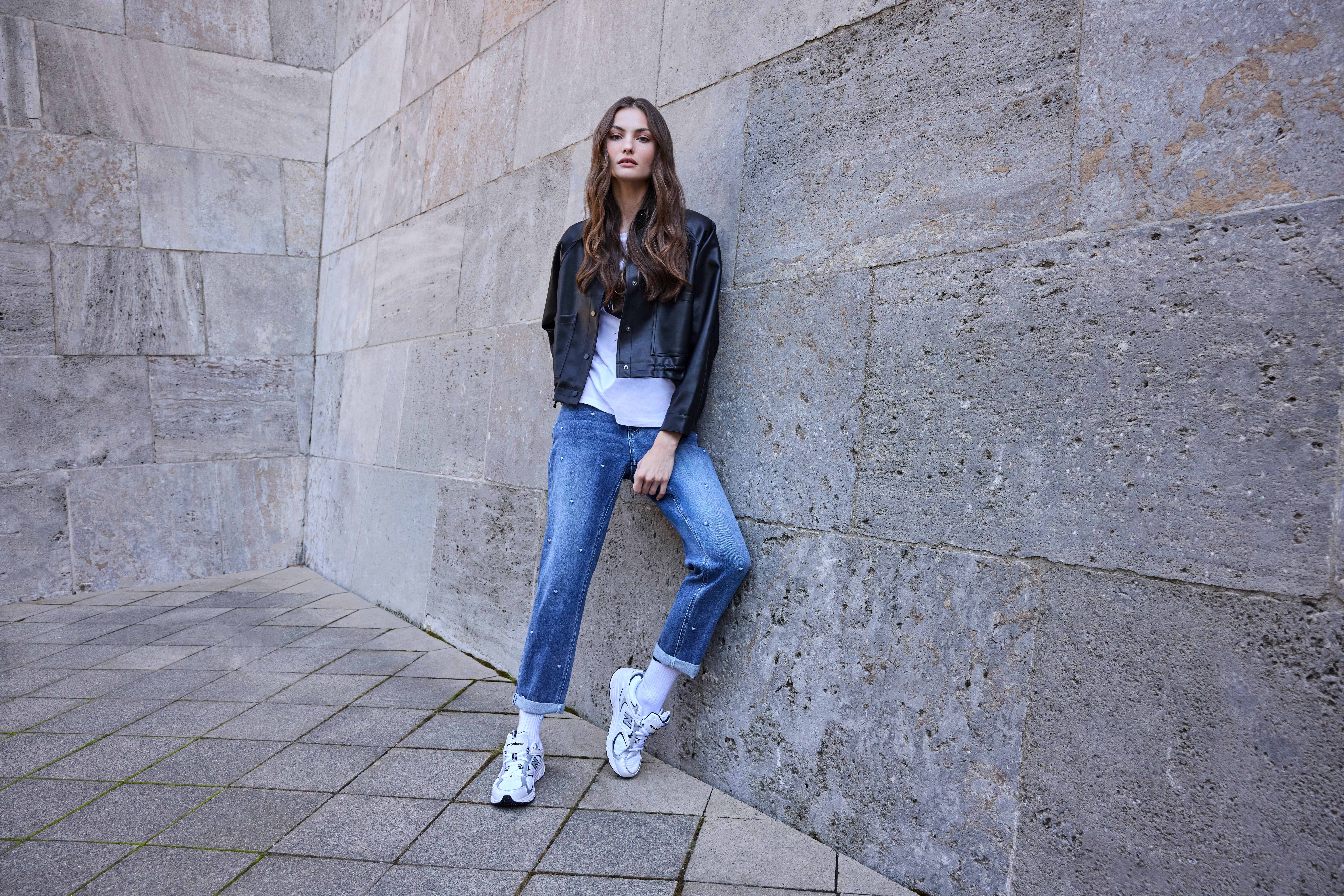 Model in Lederjacke und Perlen-Jeans mit weißen New-Balance-Sneakern vor Steinwand