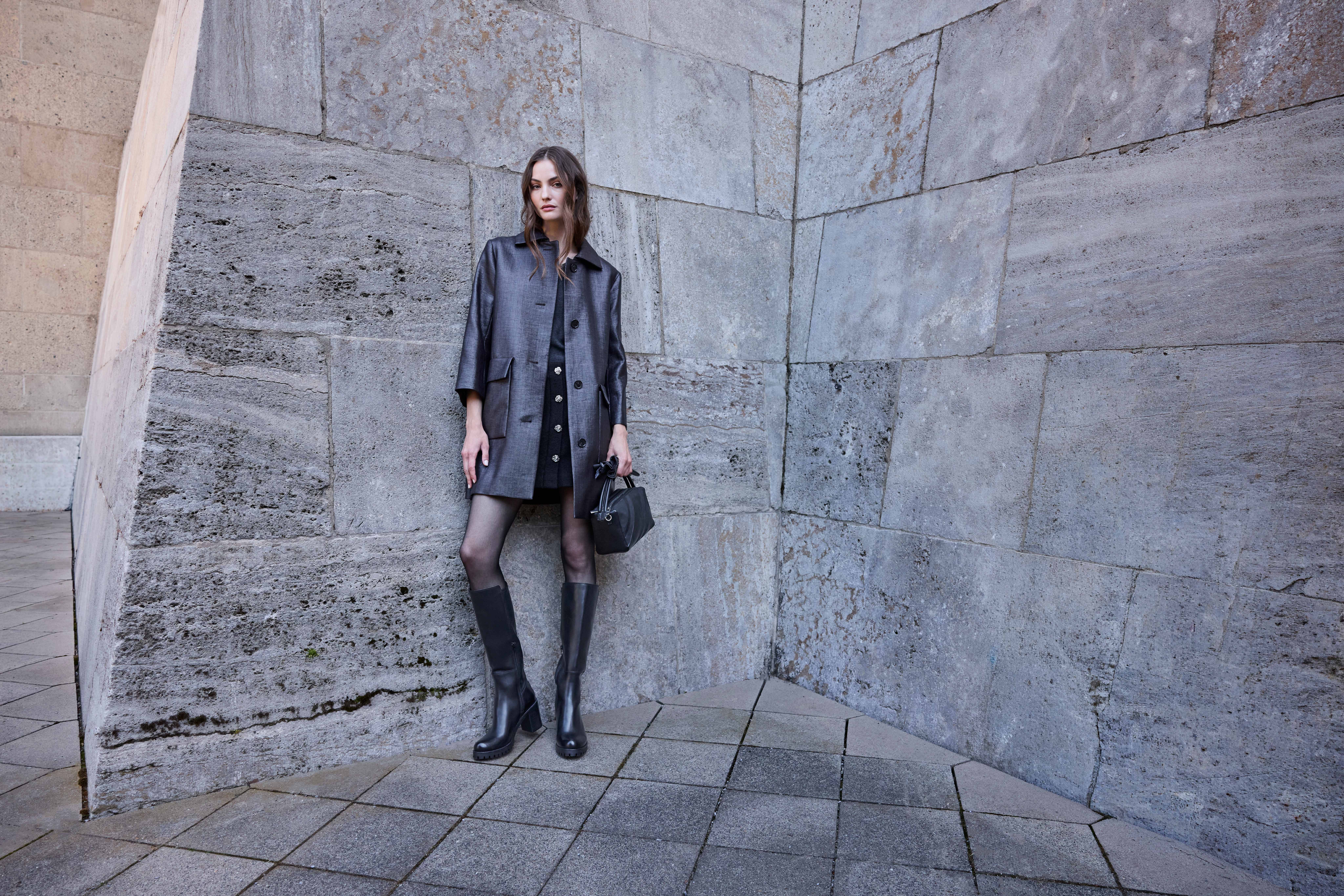 Frau in grauem Mantel und Kleid vor Steinwand kombiniert schwarze Kniestiefel mit Blockabsatz und Minitasche