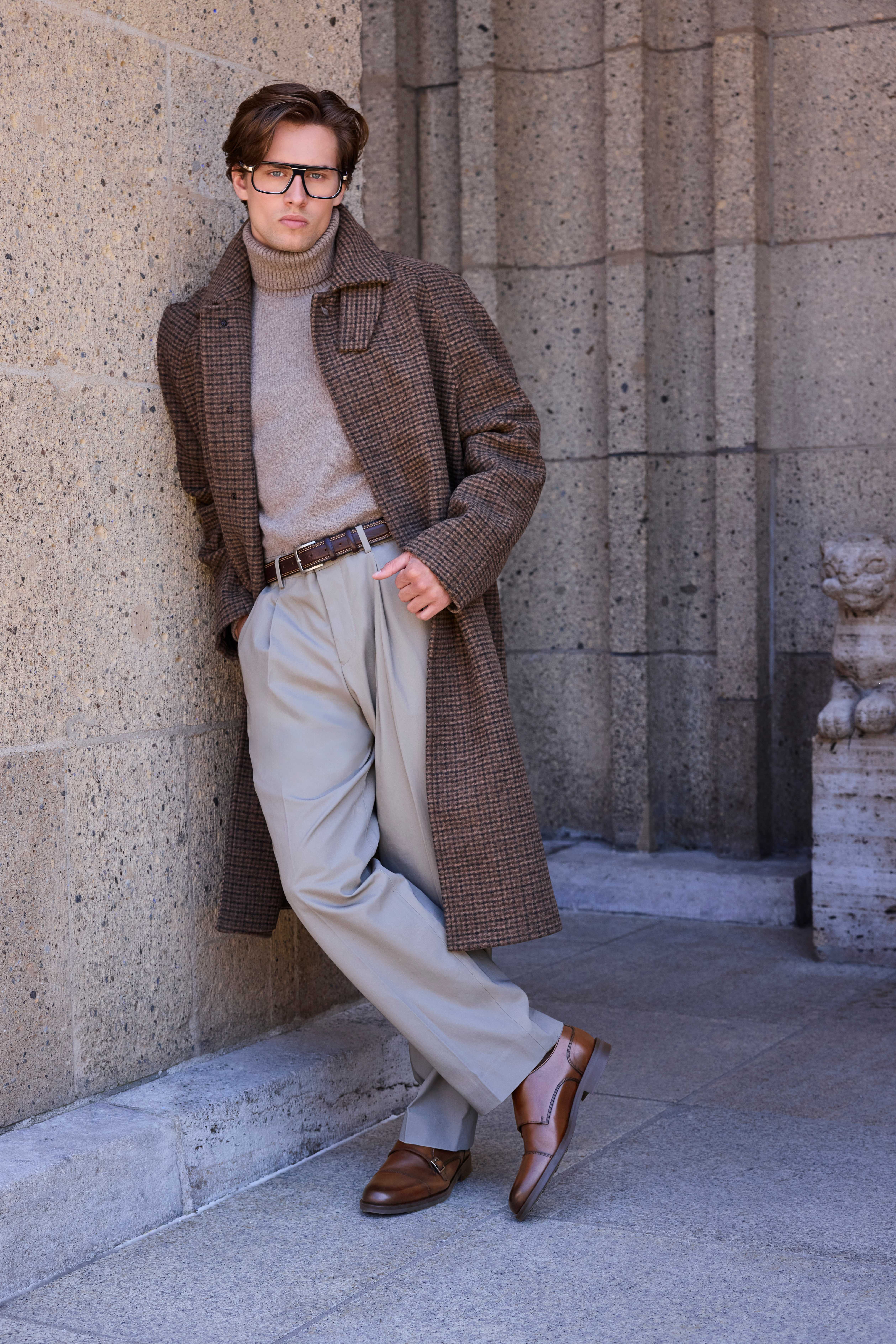 Ein Mann posiert mit einem elegantem Outfit aus einer Anzugshose, einem Rollkragenpullover, einem Mantel und glänzenden brauen Schuhen