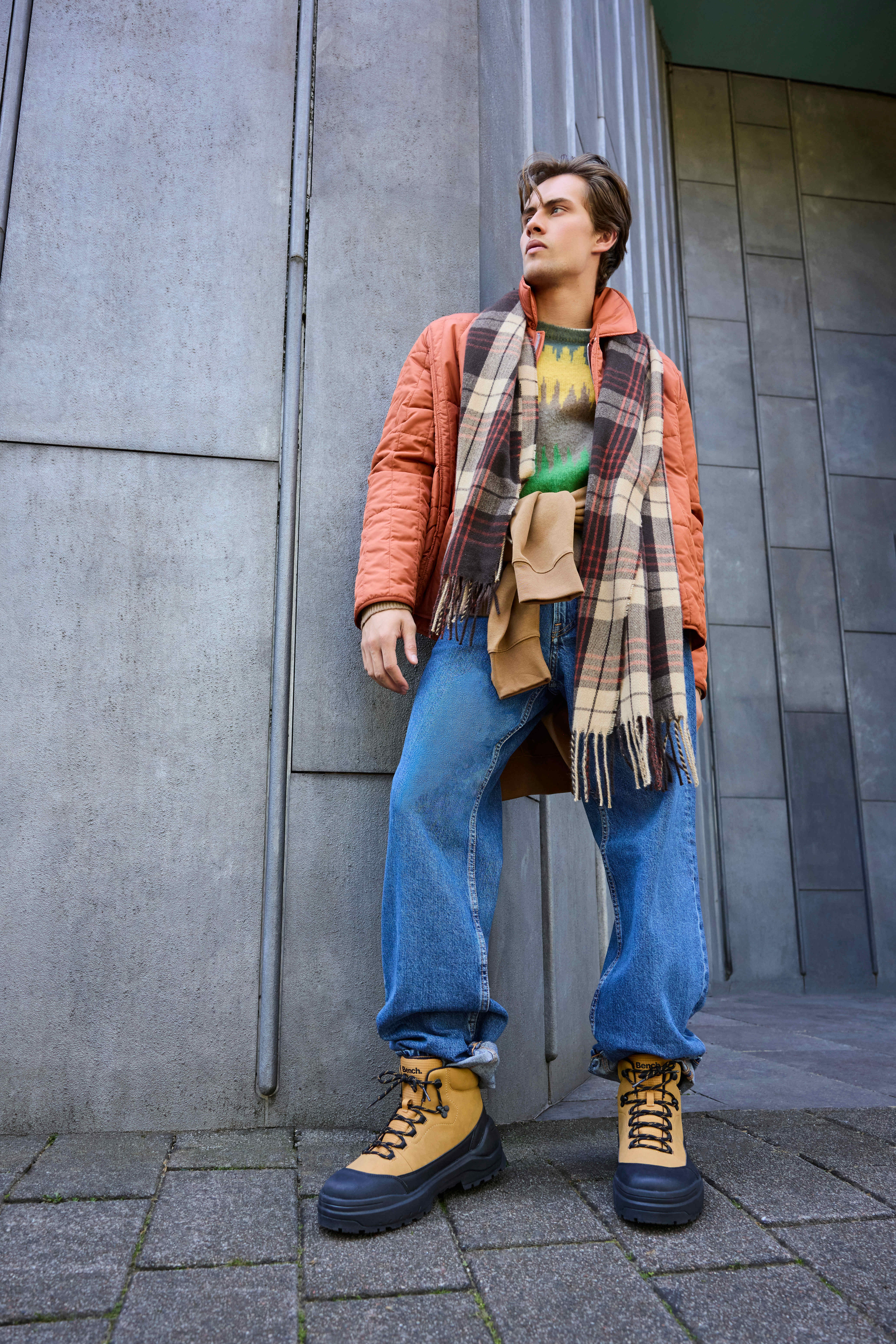 Mann in Layering-Outfit, lehnt an Wand, trägt rosa Jacke mit blauer Jeans und schweren Stiefeln