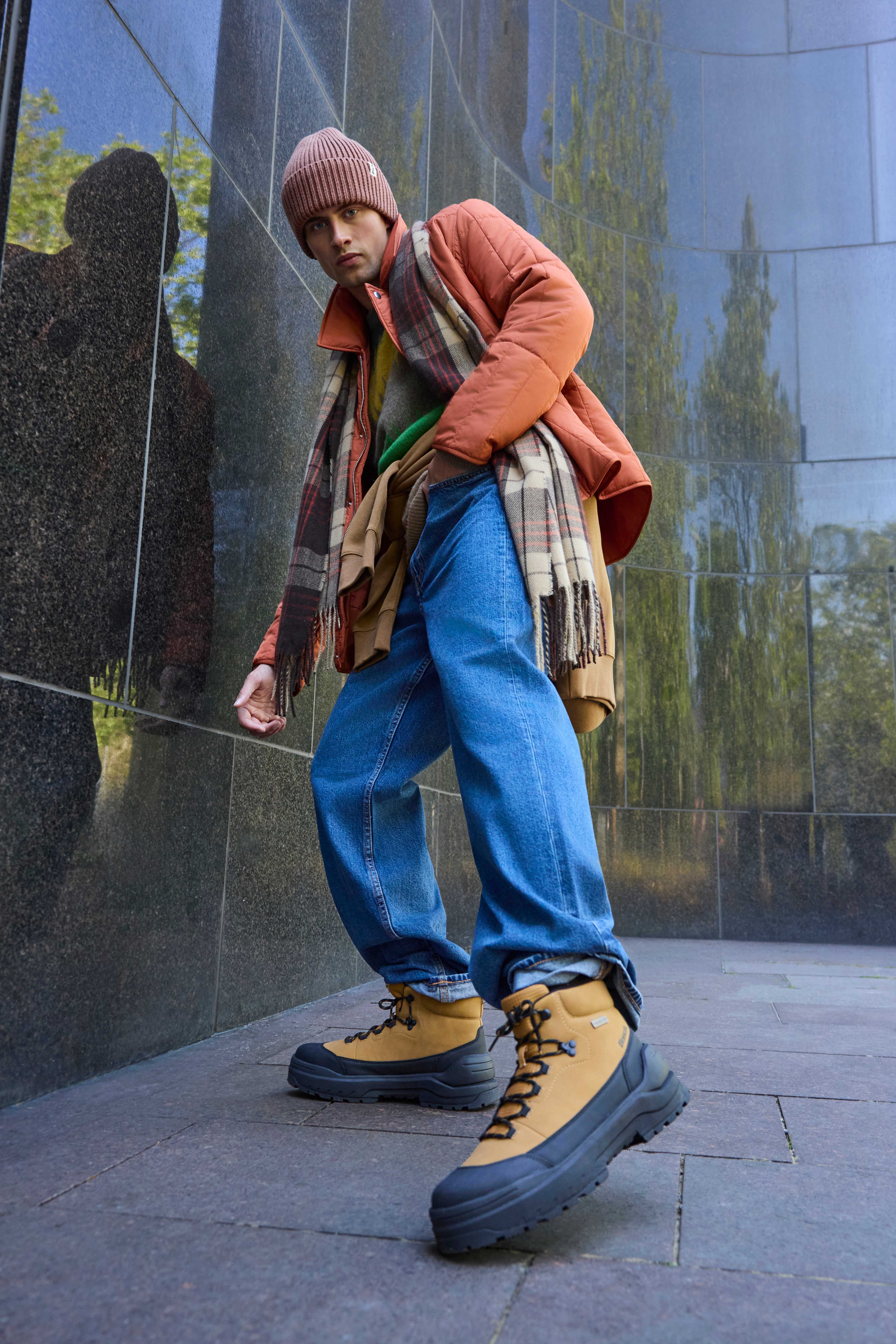 Ein Mann trägt beige Boots, mit einer Jeans, einer orangenen Jacke und Mütze 