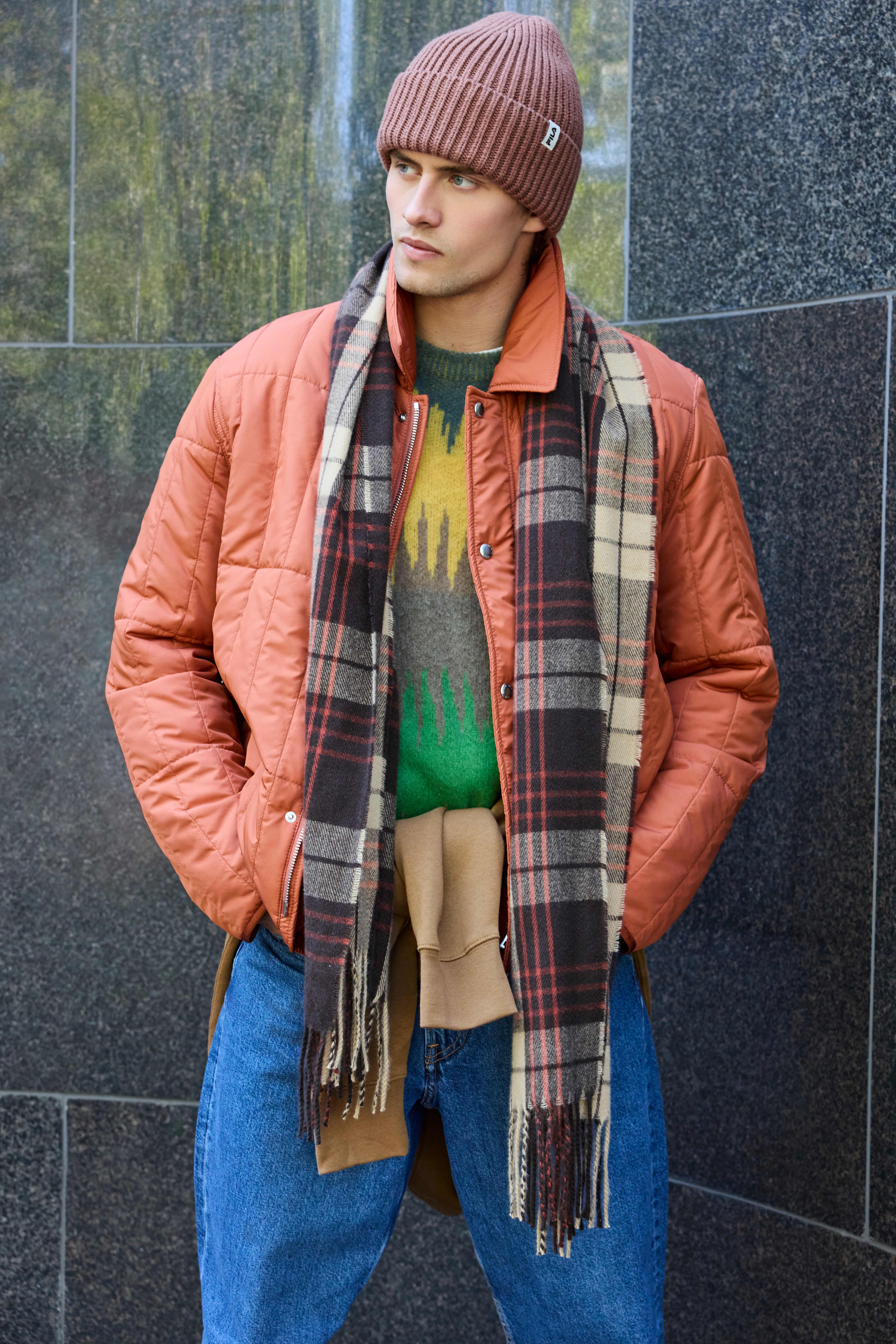Mann trägt eine braune Mütze, eine orangefarbene Steppjacke, einen karierten Schal und blaue Jeans