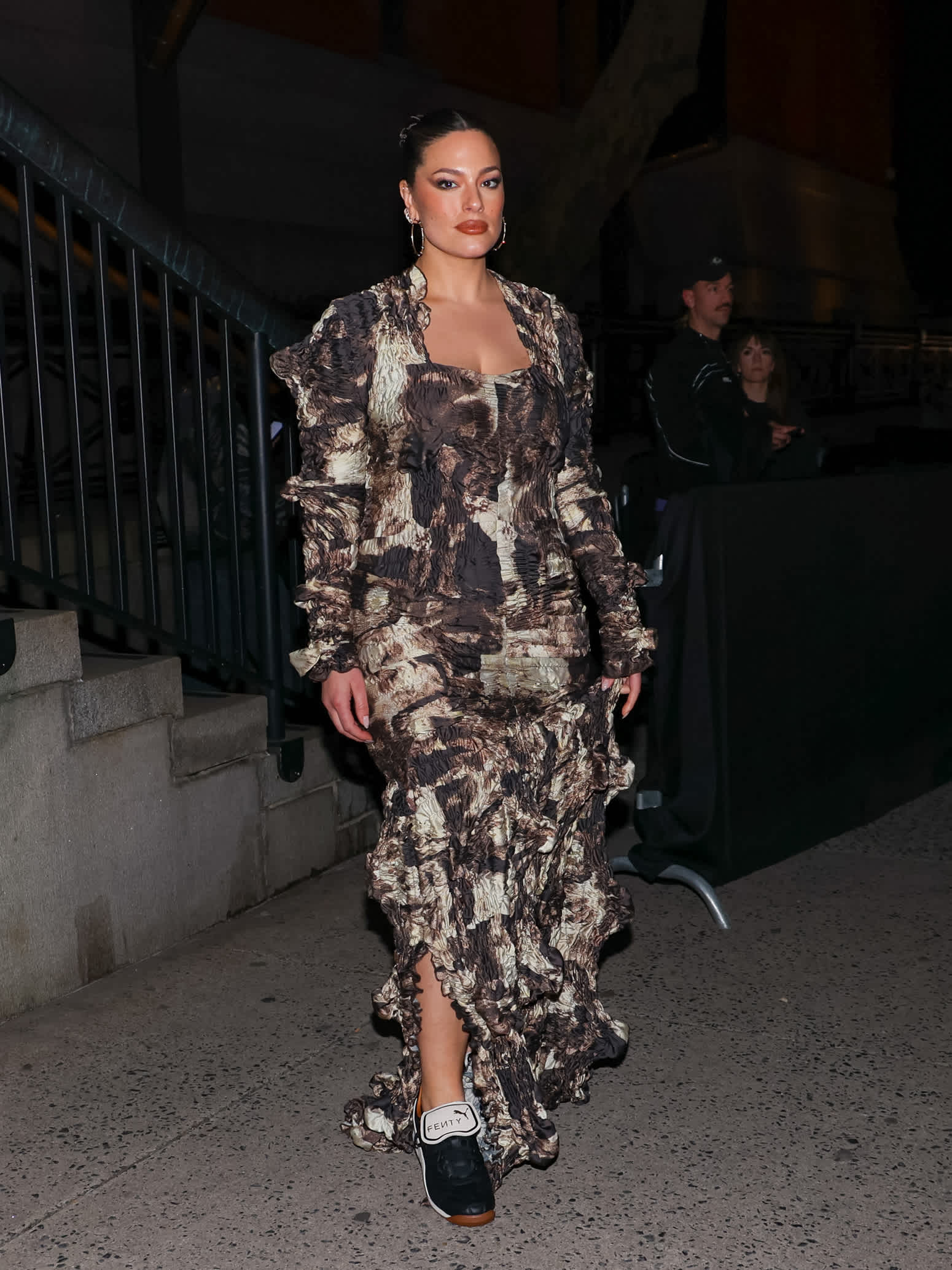 Model Ashley Graham in bodenlangem Kleid mit abstraktem braun-beigen Muster, kombiniert mit Fenty-Sneakern von PUMA 
