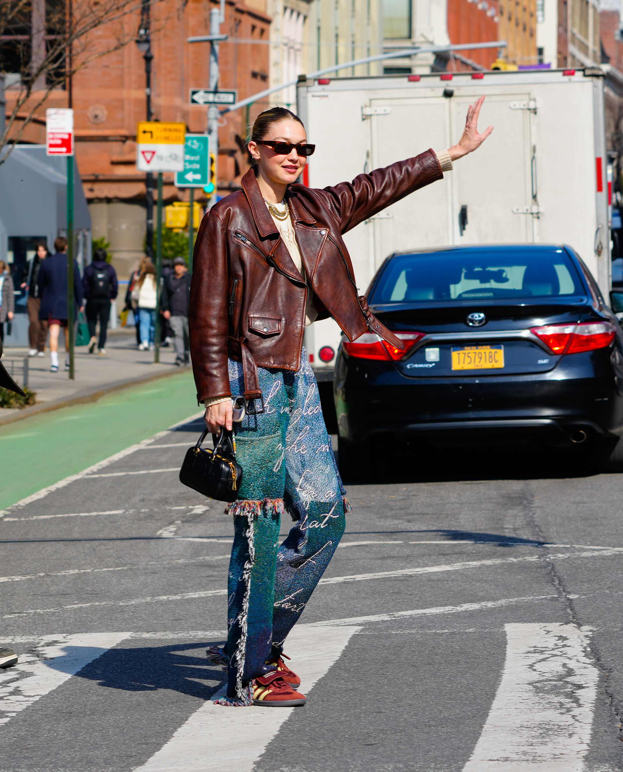 Model Gigi Hadid, trägt braune Lederjacke, bedruckte Jeans und markant rote Mom-Sneaker, auf einer Straße stehend