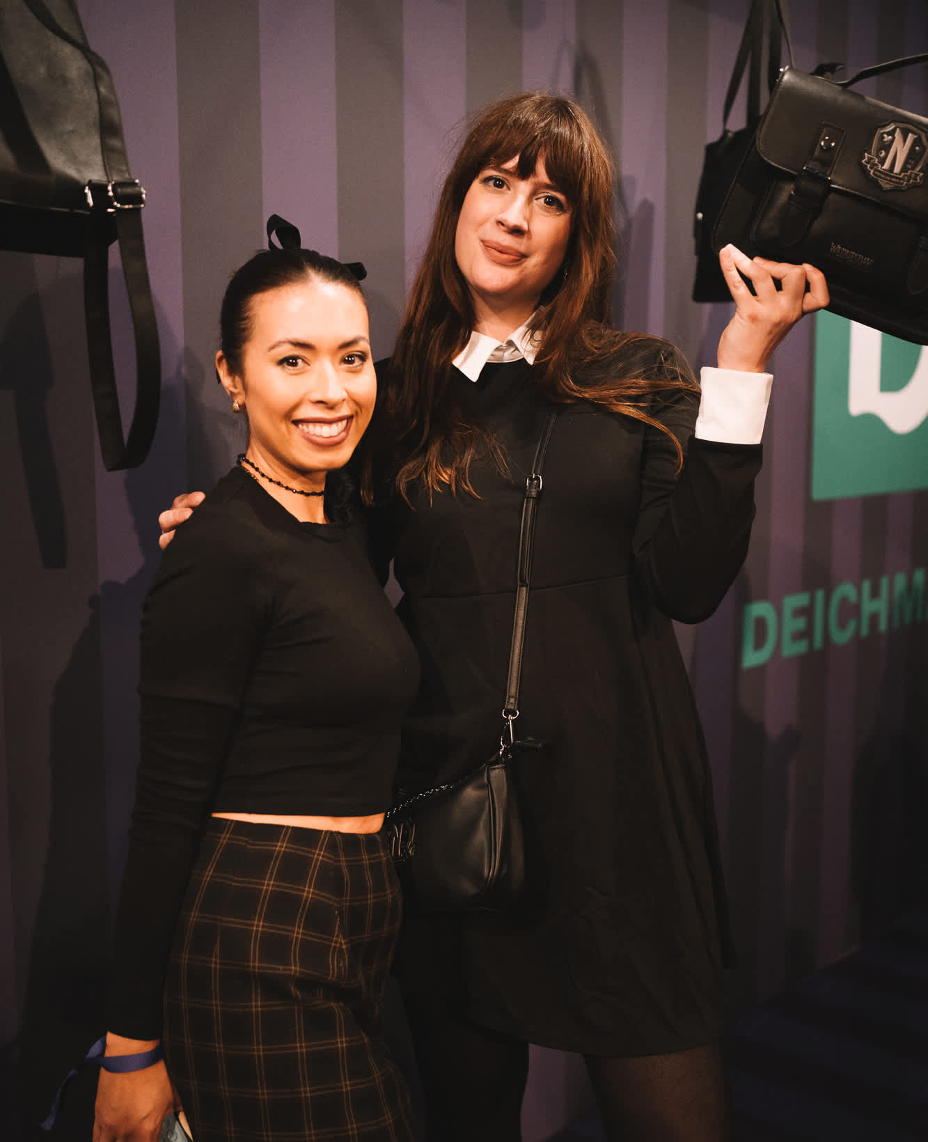 Zwei Frauen lächelnd bei einem Event, rechte Frau hält schwarze Handtasche hoch, grünes Deichmann-Logo im Hintergrund