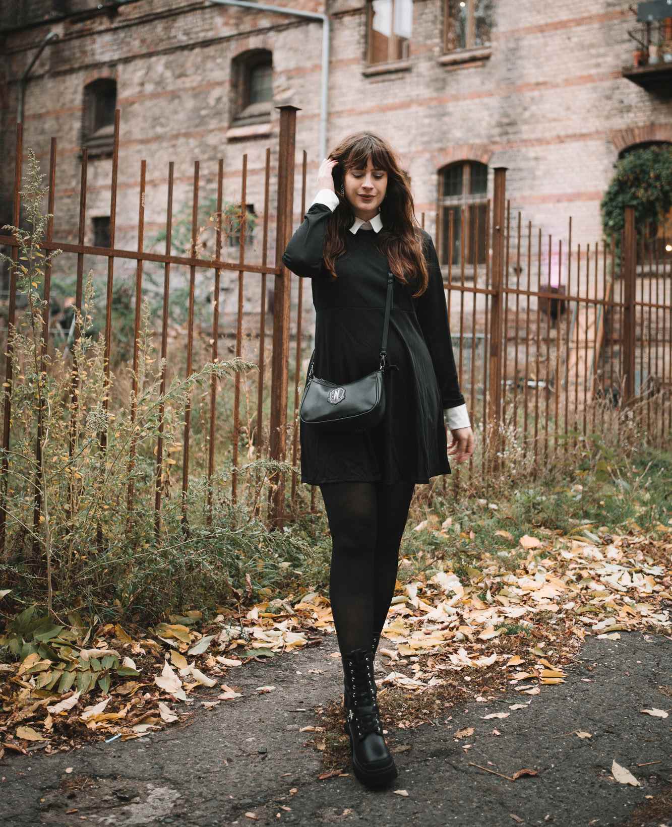 Frau in schwarzem Kleid mit weißem Kragen, schwarzer Strumpfhose und schwarzen Schnürstiefeln, gekleidet im Wednesday Addams-Stil, trägt eine Umhängetasche mit Wappen-Logo.
