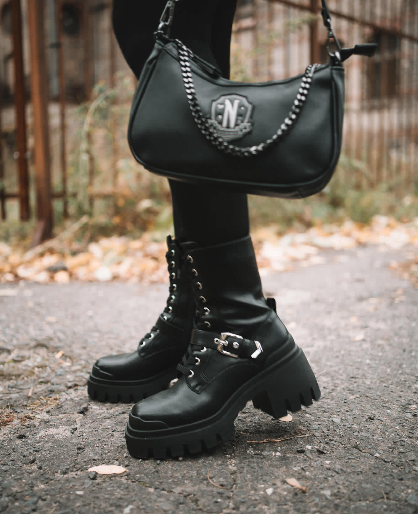 Nahaufnahme einer Person mit schwarzen Schnürstiefeln, die eine schwarze Umhängetasche mit Wappen-Logo und Kettendetail hält