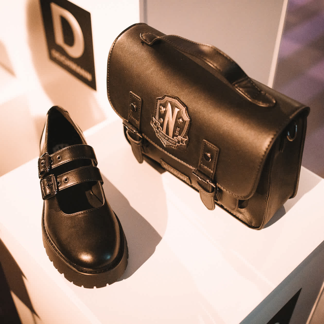 Nahaufnahme eines schwarzen Schuhs und einer passenden schwarzen Tasche mit einem Wappen-Logo, ausgestellt auf einem weißen Podest, mit Deichmann-Logo im Hintergrund