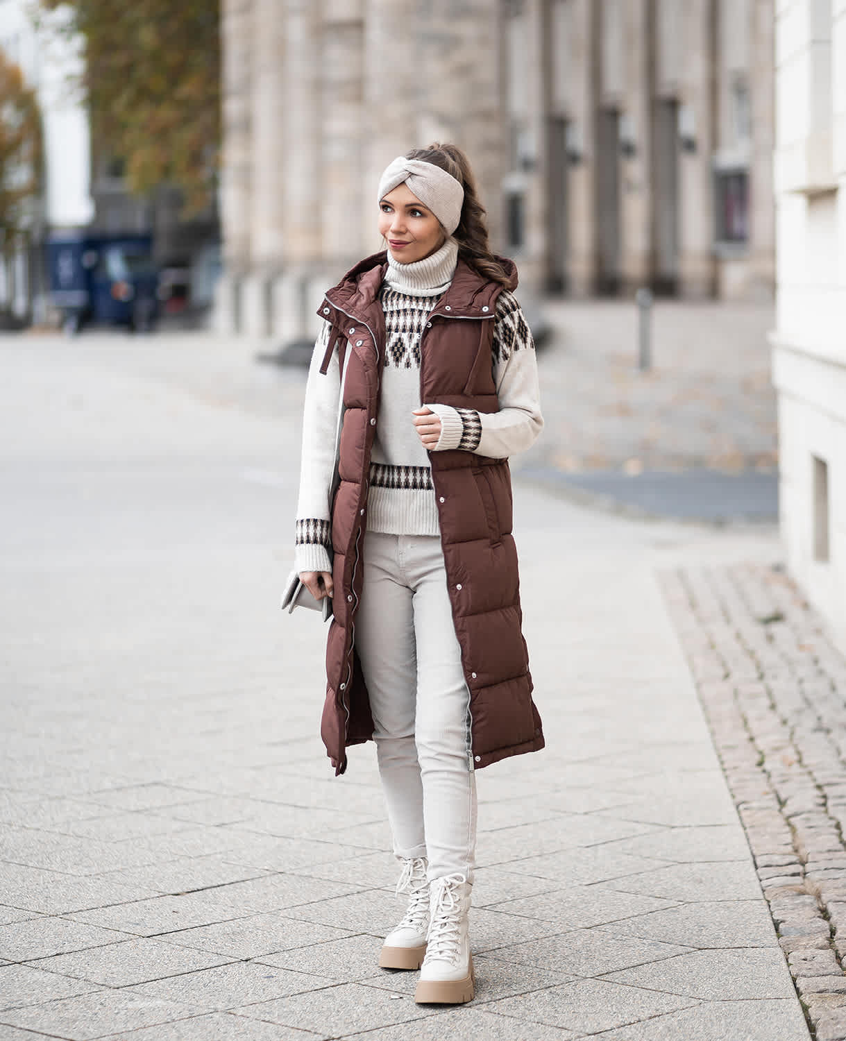 Frau in winterlichem Look mit brauner Weste, Norwegerpullover und beigen Schnürboots, spazierend in urbaner Umgebung