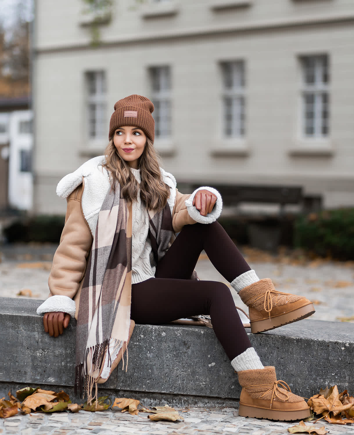 Frau in kuscheliger Shearling-Jacke in Beige, Strickmütze, Schal und Fake Fur-Boots, entspannt sitzend im Freien