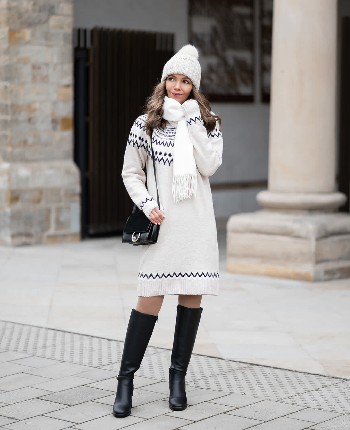 Frau in weißem Strickkleid, Strickmütze und Schal, kombiniert mit schwarzen Stiefeln und Handtasche